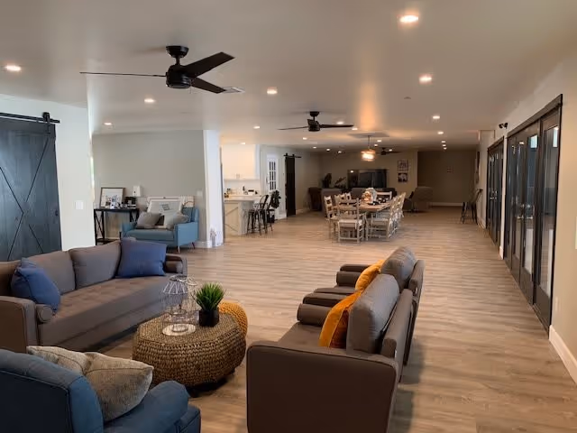 Spacious open communal living area with sofas, armchairs, dining tables, ceiling fans, and large glass doors.