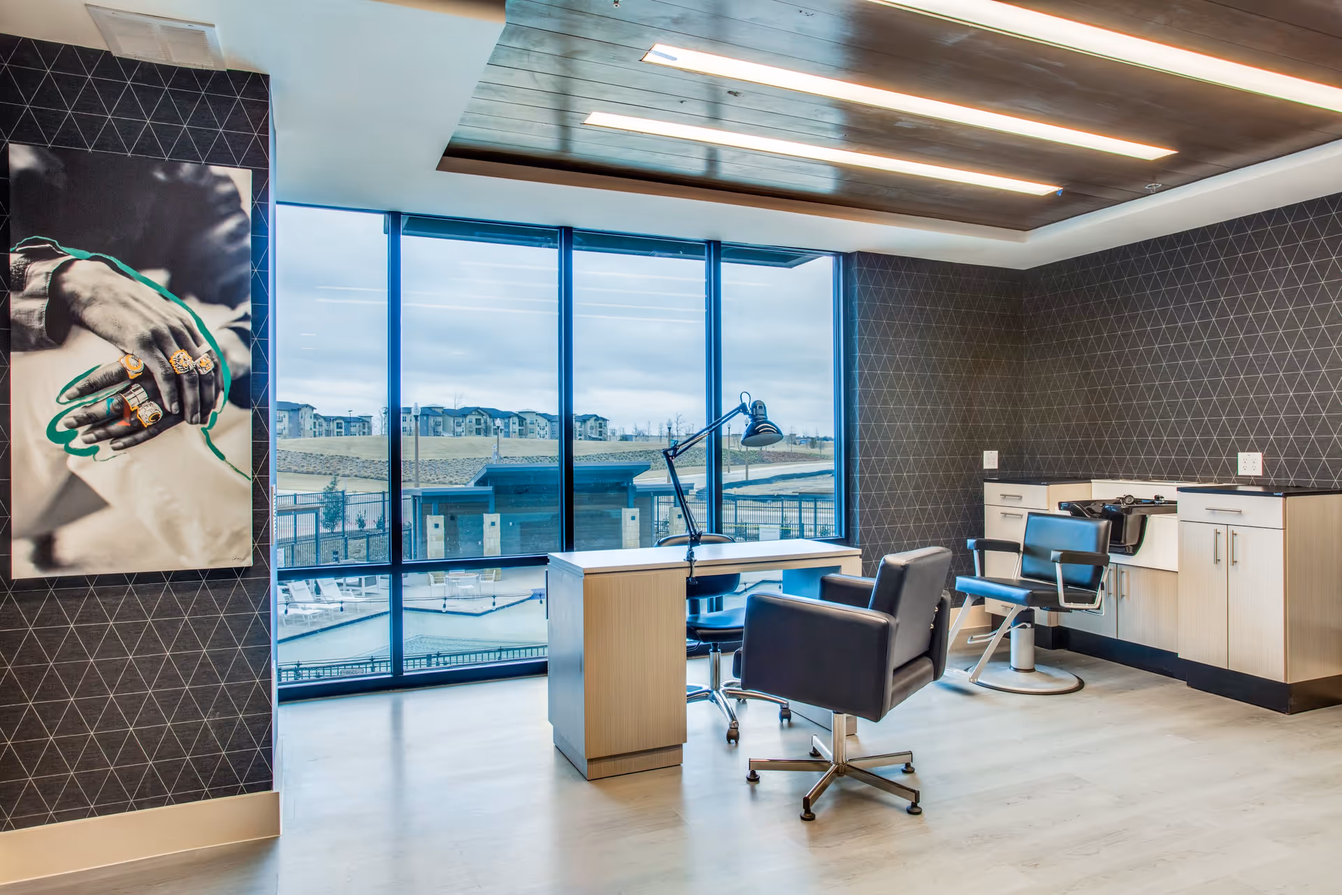 A modern salon area inside a senior living facility with two black salon chairs, a desk with a lamp, and cabinetry with a hair washing station. Large floor-to-ceiling windows provide a view of an outdoor pool area and buildings in the distance. The walls have a geometric pattern wallpaper and a large black and white photo of hands with rings is displayed on one wall.