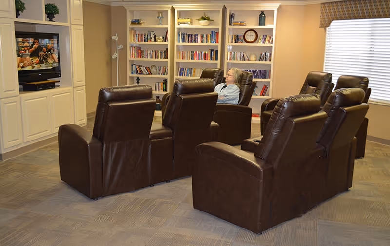 A cozy common room with six brown leather recliners arranged in two rows facing a television mounted in a white built-in cabinet. A woman is sitting in one of the recliners watching the TV. Behind the chairs, there are built-in bookshelves filled with books and decorative items. A window with closed blinds and a valance is on the right wall.