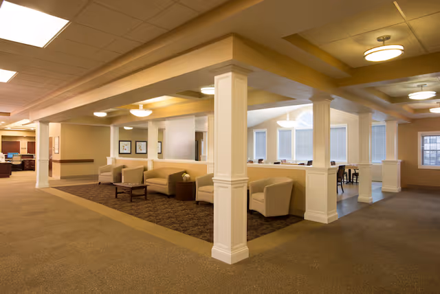A spacious and well-lit interior common area with beige walls and carpeted floors. The area features several beige armchairs and sofas arranged around small wooden tables. White columns and half walls separate the seating area from an adjacent room with large windows and additional seating. Ceiling lights provide warm illumination throughout the space.