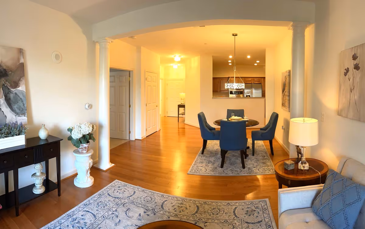 Interior view of a retirement community living space featuring a small dining area with a round table and four blue upholstered chairs on a patterned rug. The room has hardwood floors, white walls, decorative columns, and a kitchen area visible in the background. There is a side table with a lamp and a beige armchair with a blue cushion, as well as a black console table with decorative items and a potted plant.