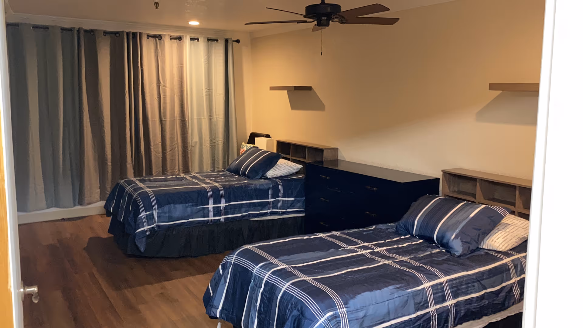 A bedroom with two twin beds covered in blue plaid bedding. Each bed has pillows and a small wooden headboard with cubby storage. There is a dark dresser between the beds and two floating shelves on the wall above it. The room has wood flooring, a ceiling fan with lights, and a window covered by gray curtains.