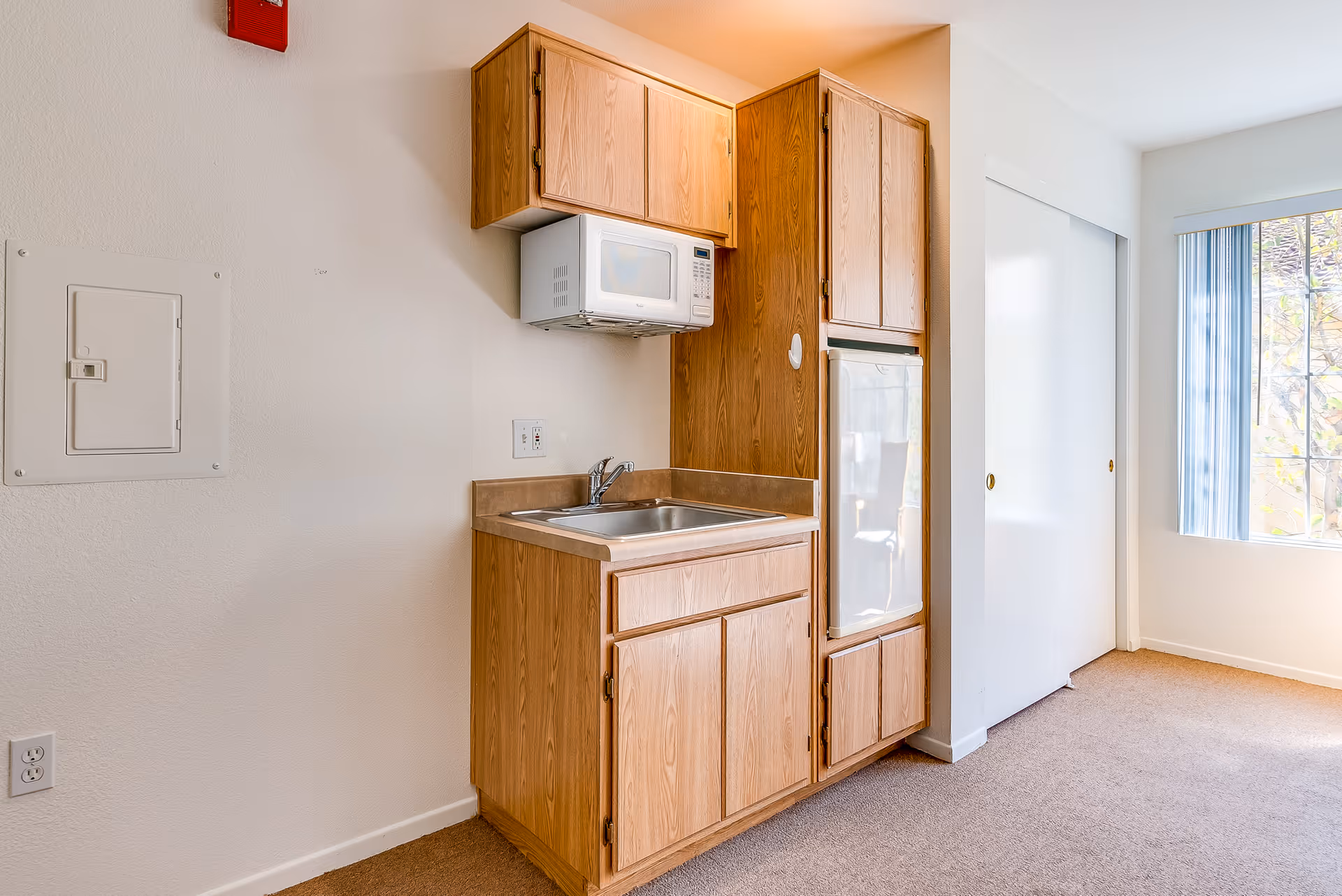 A compact kitchenette with wood cabinets, a sink, an over-the-sink microwave, and a small refrigerator beside a window and closet.