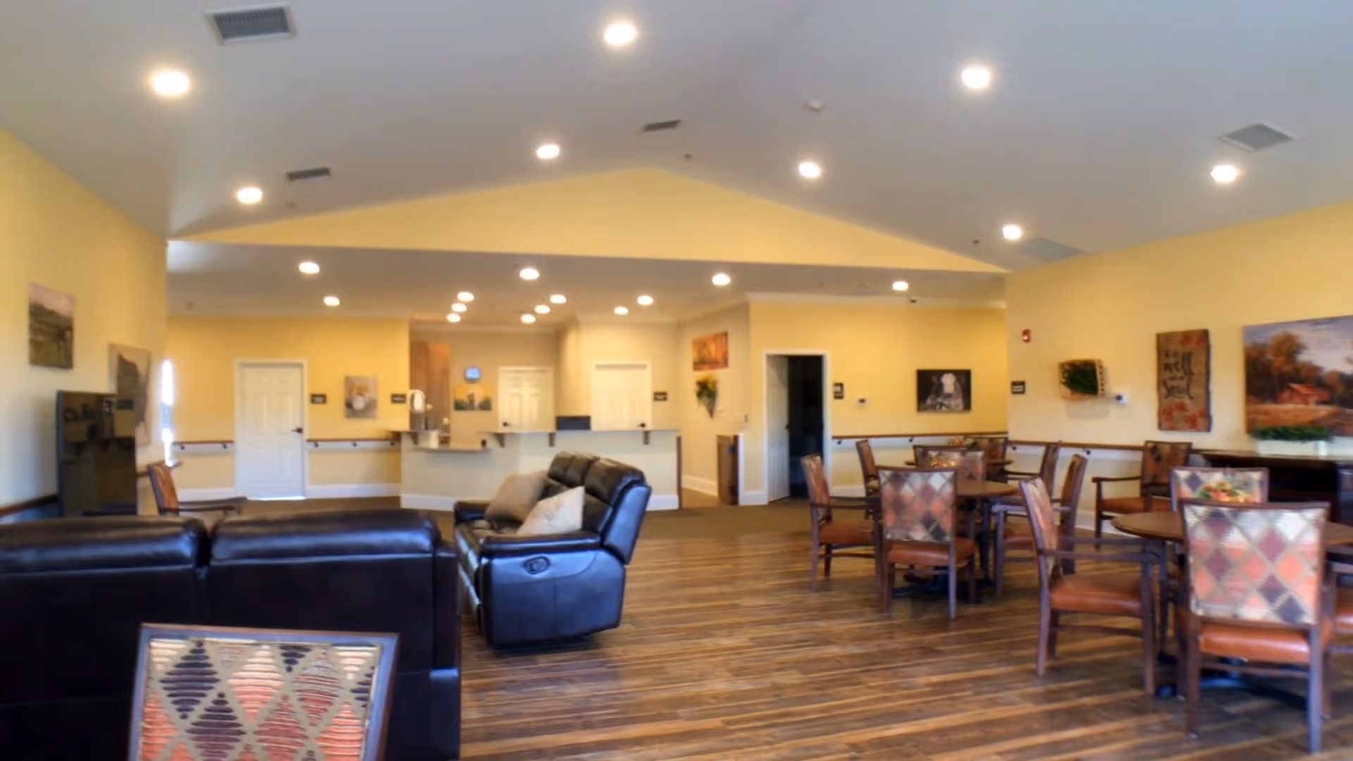 Interior view of a senior living facility common area with yellow walls, wood flooring, several round tables with chairs, and a seating area with leather sofas. The ceiling has recessed lighting, and there are paintings and decorations on the walls.