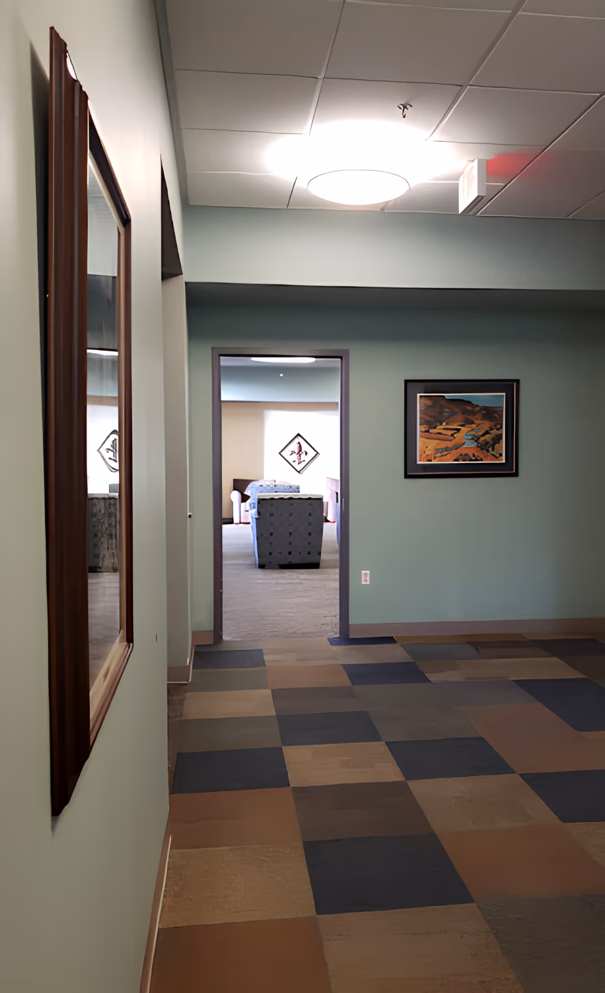 A pastel-green hallway with patterned tile flooring leading to a doorway revealing a seating area, with a mirror on the left and framed artwork on the right.