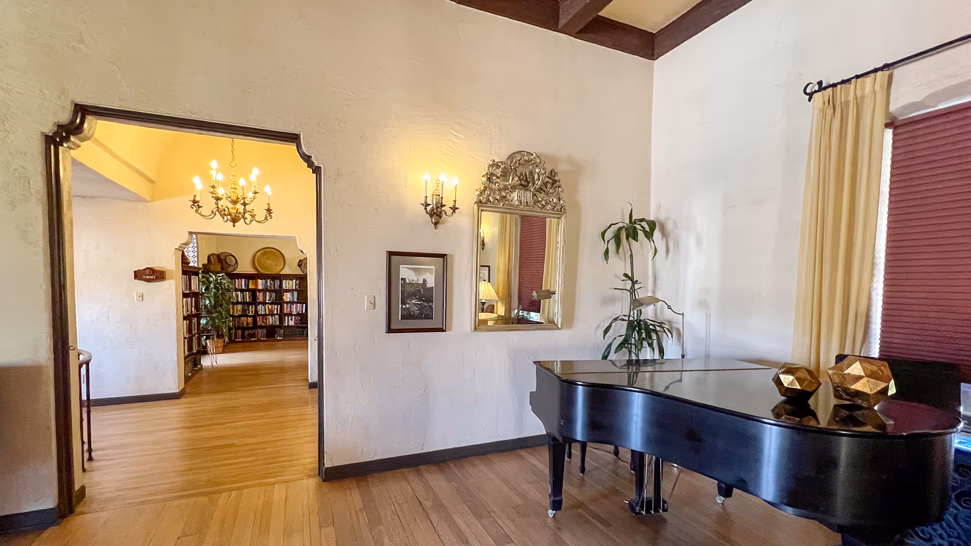 Interior view of a senior living facility room featuring a black grand piano on the right, a decorative mirror and framed picture on the wall, a potted plant, and an open doorway leading to a room with bookshelves and a chandelier.
