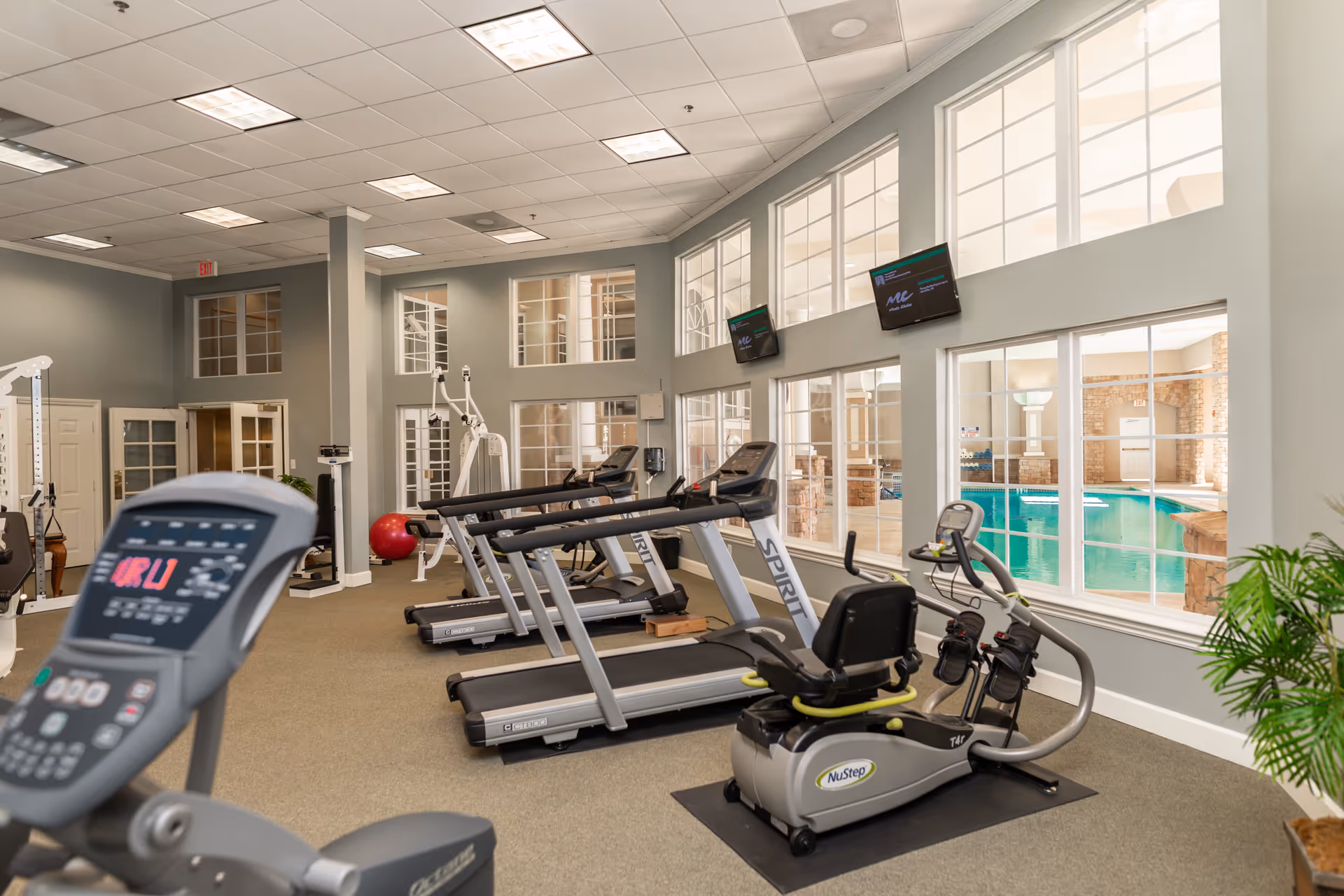 Interior view of a fitness center with several treadmills, an exercise bike, and other gym equipment. Large windows on the right side overlook an indoor swimming pool. The room has high ceilings with recessed lighting and light gray walls.