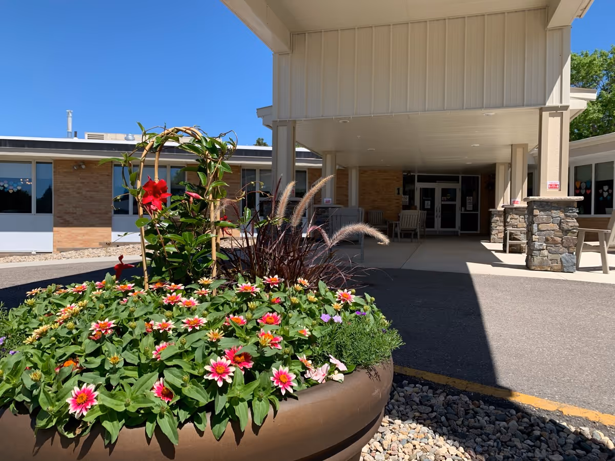 Entrance of Glenwood Retirement Village with a large flower pot filled with colorful flowers in the foreground. The building has a covered entryway supported by columns with stone bases, and large windows on either side. The sky is clear and blue.