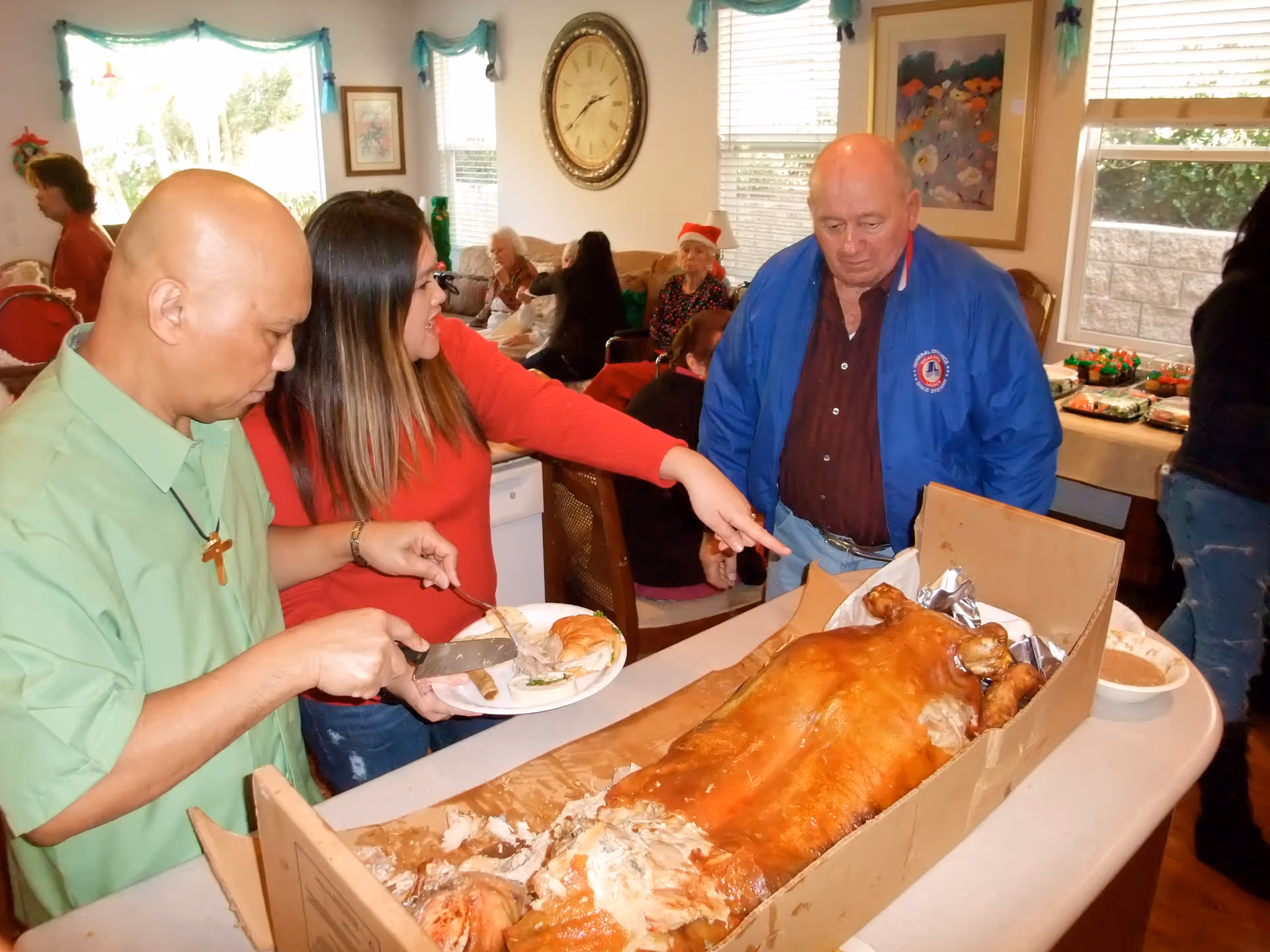 Three people serving and looking at a large roasted pig on a table inside a room decorated for a gathering. One person is carving the pig, another is holding a plate, and the third is pointing at the pig. In the background, several elderly people are seated on couches and chairs near windows with blinds and holiday decorations.