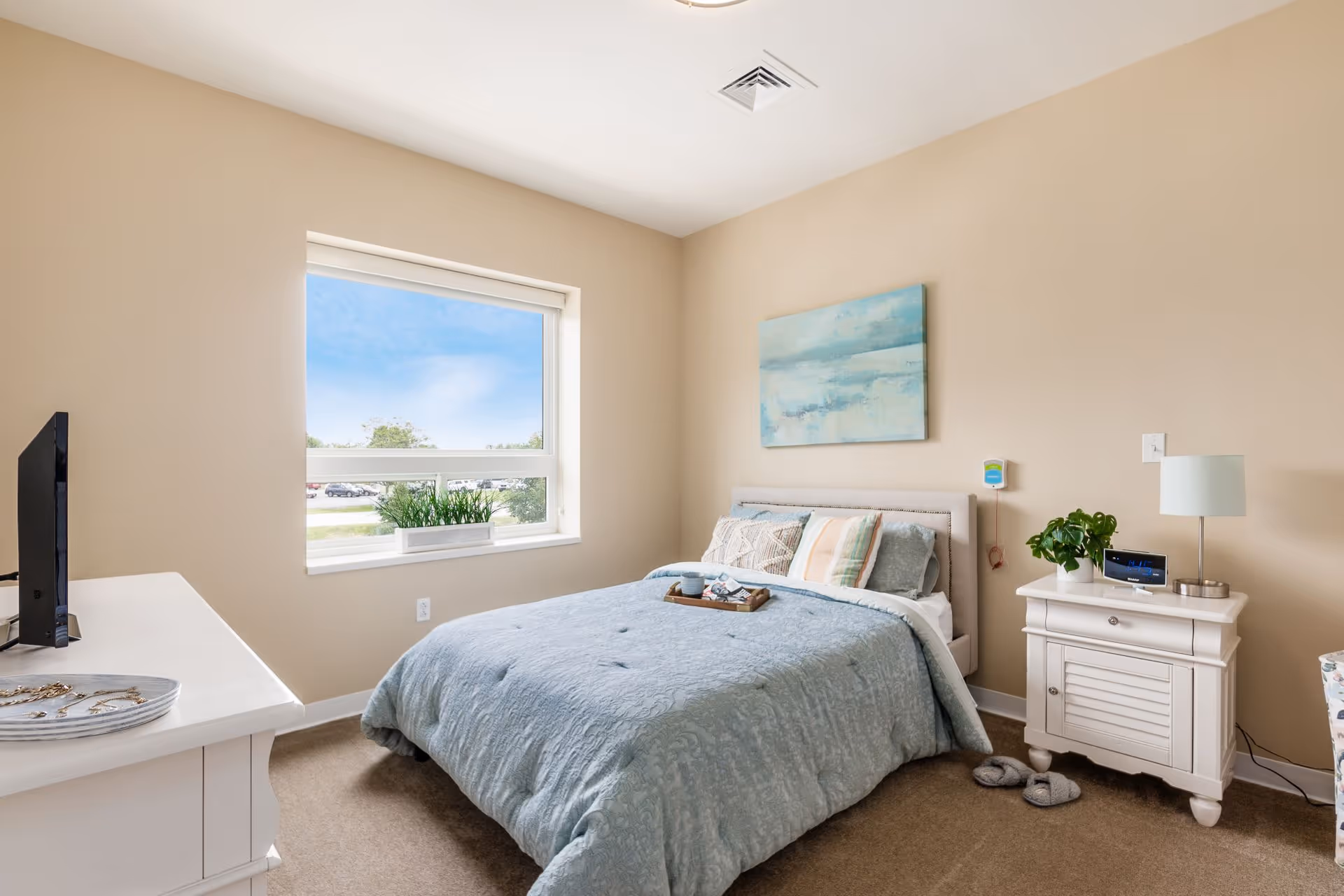A cozy bedroom with a neatly made bed covered in a light blue quilt and several pillows. A small tray with a cup and a book rests on the bed. There is a white nightstand beside the bed with a lamp, a small plant, and a digital clock. A window with a view of a parking lot and trees lets in natural light. A dresser with a TV on top is visible on the left side of the room. The walls are painted beige and a blue abstract painting hangs above the bed.
