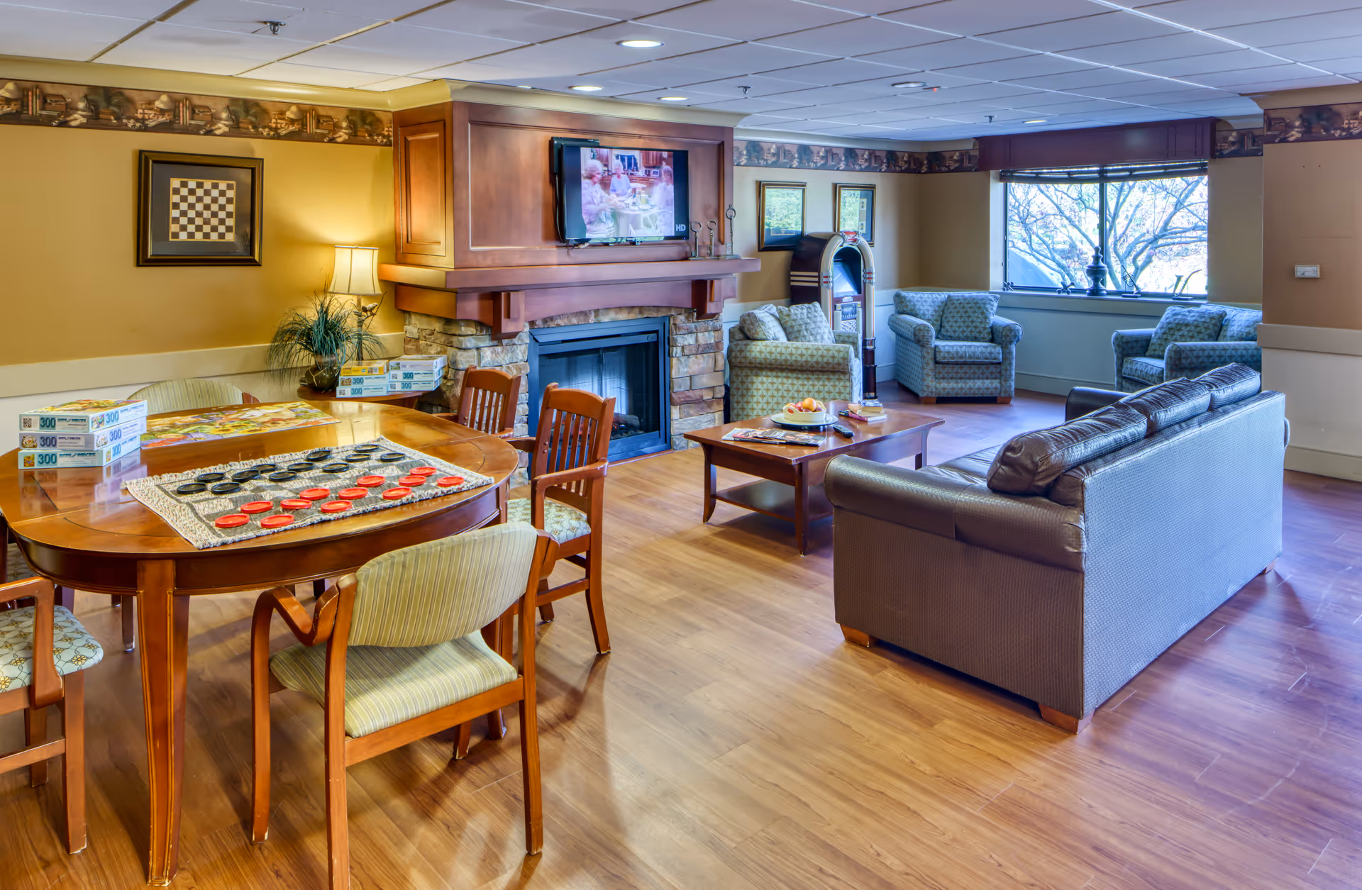 A cozy living room area with a fireplace and a mounted TV above it. There are several comfortable armchairs and a sofa arranged around a wooden coffee table. A wooden table with chairs is set up with a checkers game and puzzle boxes. A large window lets in natural light, and the room has warm yellow walls and wood flooring.