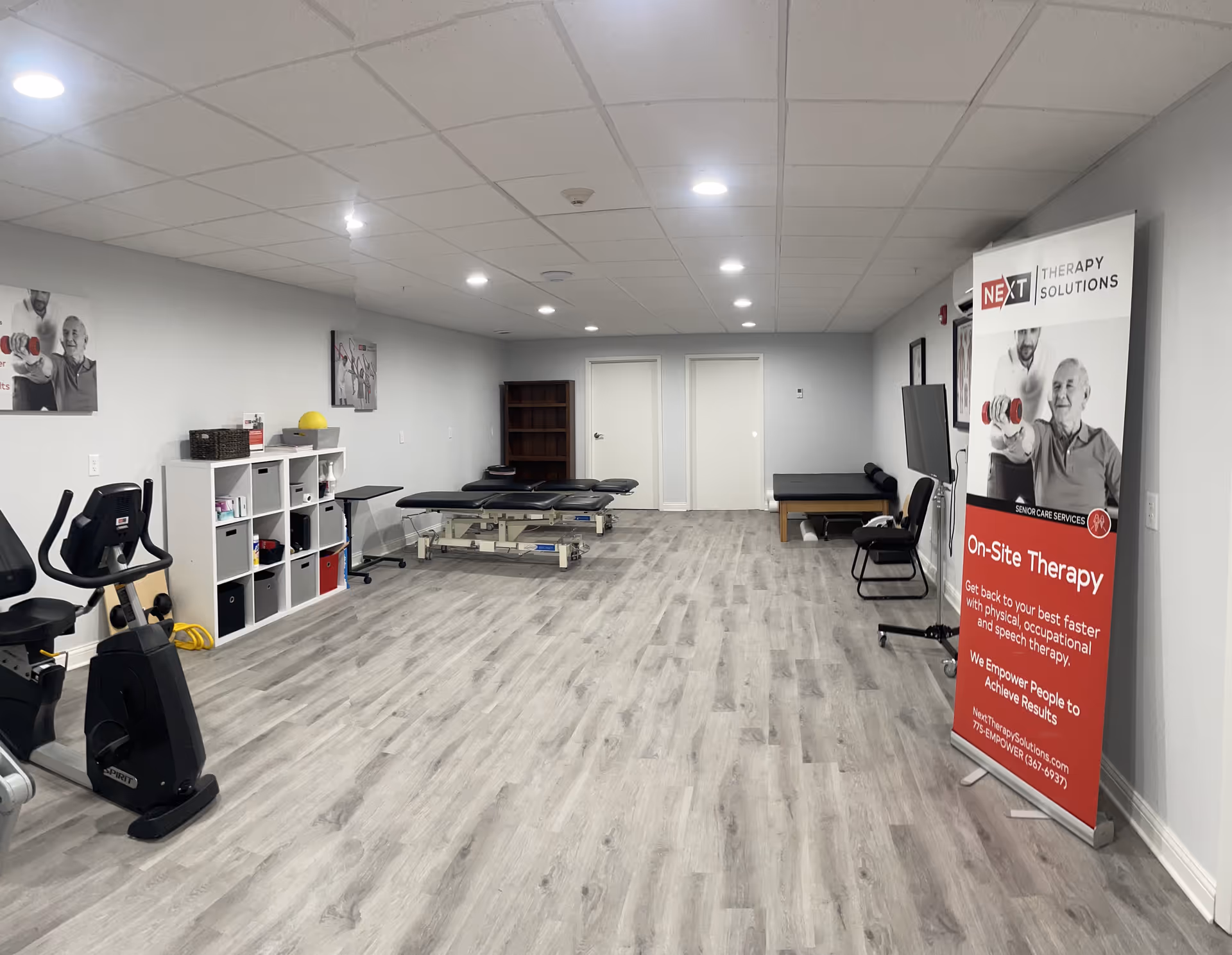 A therapy room with exercise equipment including a stationary bike, therapy tables, and storage shelves. The room has light gray walls, wood-patterned flooring, and bright ceiling lights. A large banner on the right side advertises Next Therapy Solutions offering on-site therapy services for physical, occupational, and speech therapy.