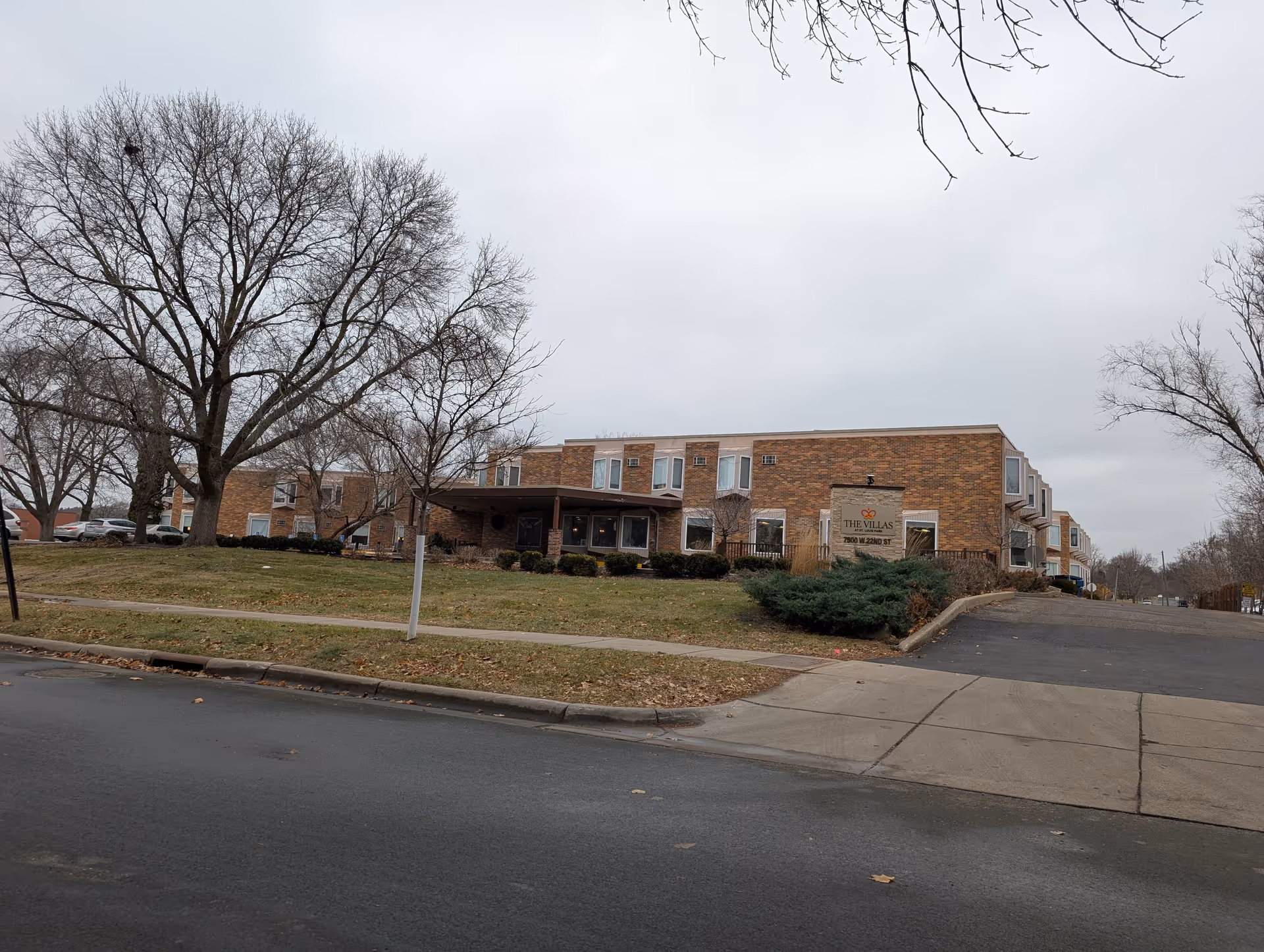 Exterior view of a brick building with a sign that reads 'The Villas at St Louis Park 7800 W 22nd St'. The building is surrounded by leafless trees and a grassy area with a sidewalk and a driveway leading up to the entrance. The sky is overcast.