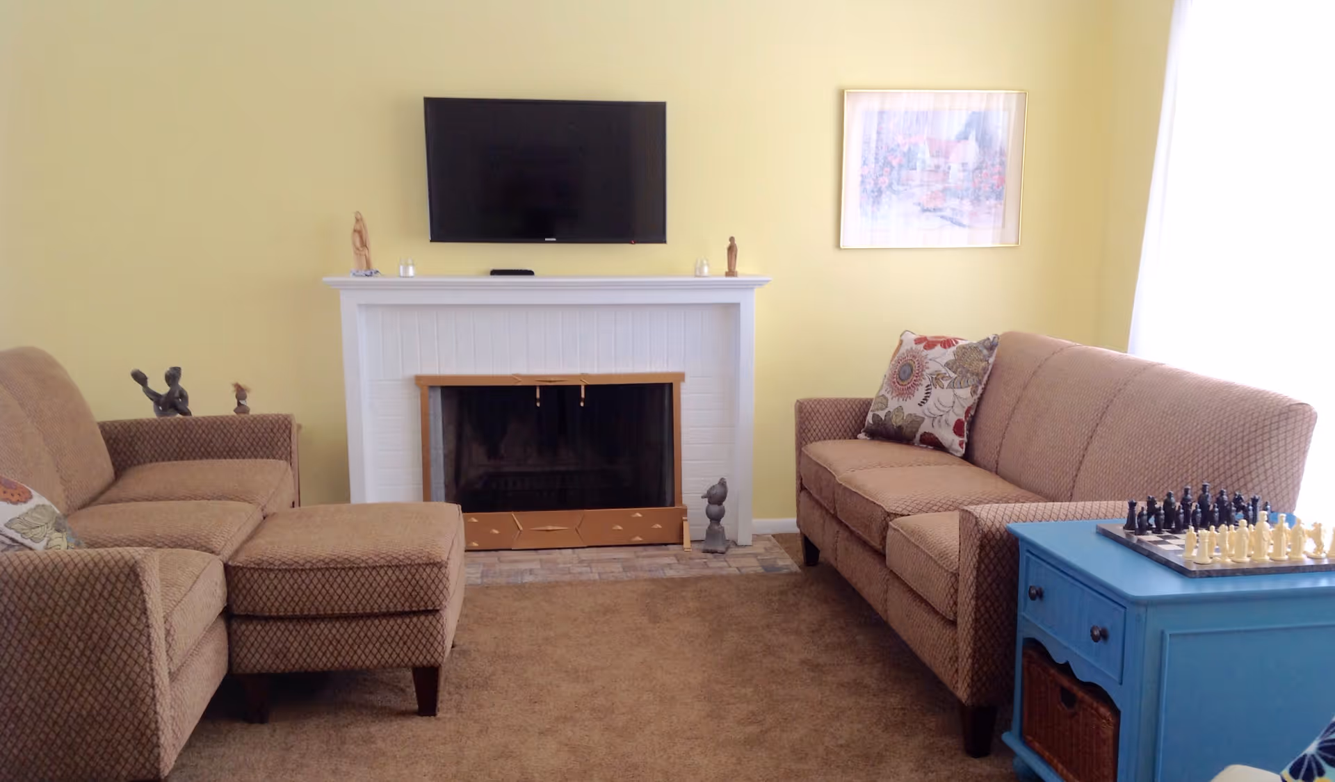 A cozy living room with two beige patterned sofas facing each other, a white fireplace with a mounted flat-screen TV above it, and a blue side table with a chessboard on top. The walls are painted yellow, and there is a framed picture hanging on the wall next to a window with white curtains.