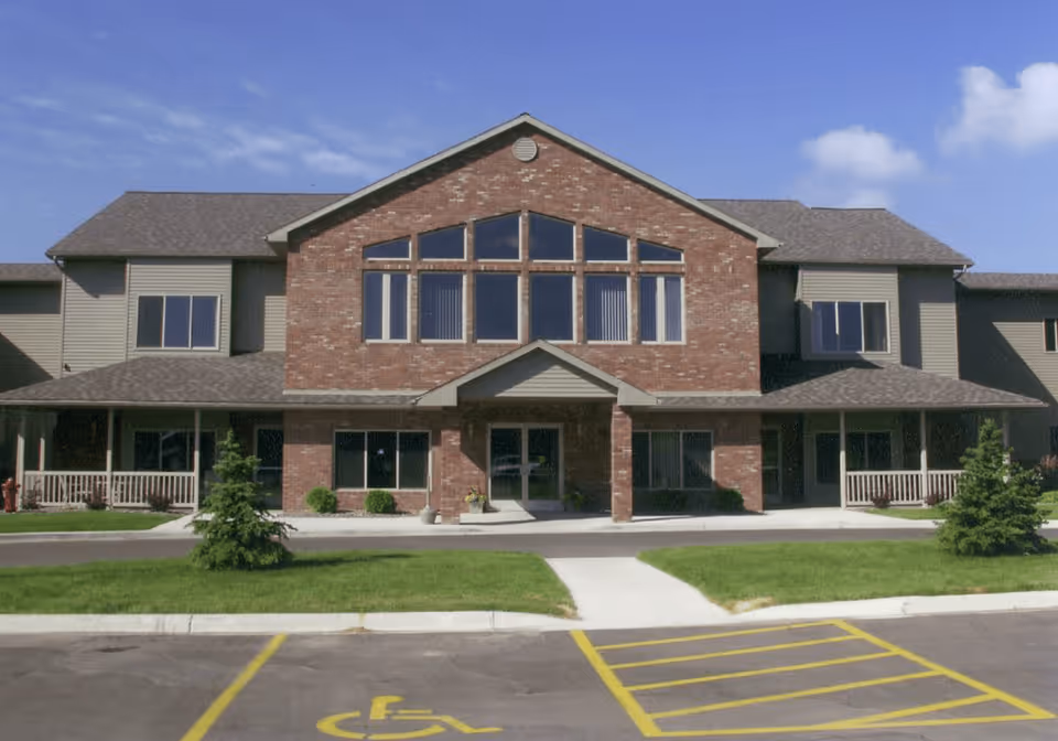 Front exterior view of South Grove Lodge Senior Living building with a brick facade, large windows, a covered entrance, green lawn, small trees, and a parking lot with a handicapped parking space in front.