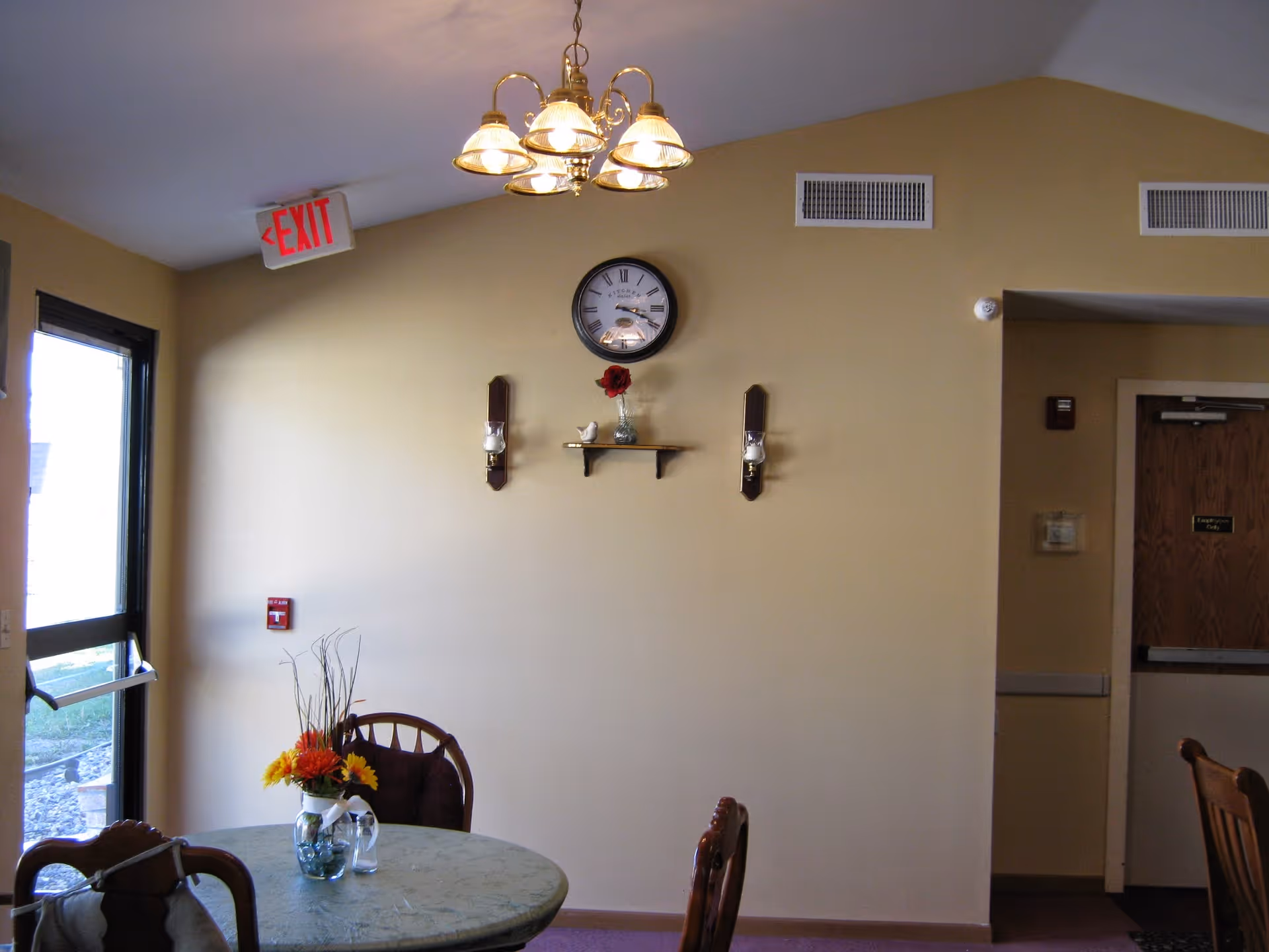 Small common dining area with a round table and flower centerpiece, wall clock, chandelier, and an exit door.