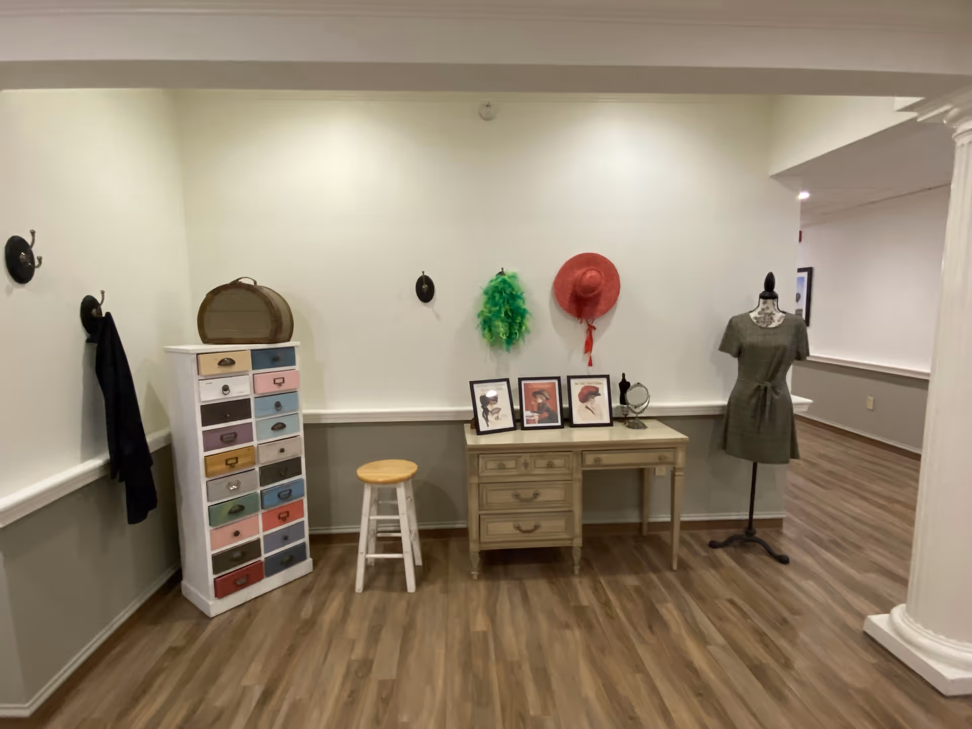 Interior activity corner with a desk, colorful drawer cabinet, mannequin in a dress, hats and framed pictures on the wall, and a stool on wood-look flooring.