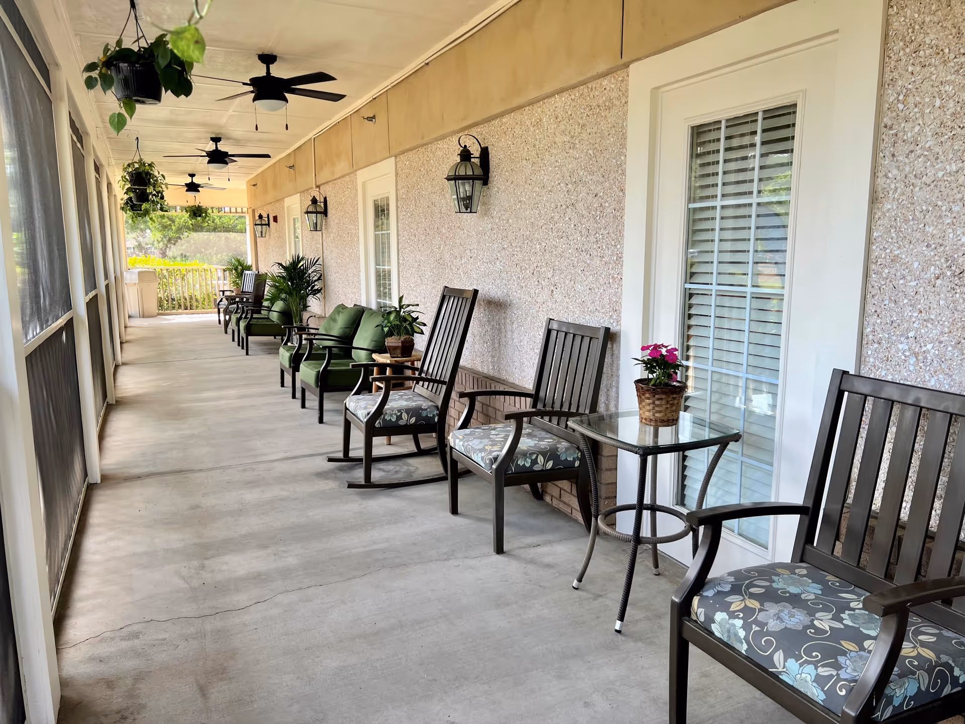 A long covered porch with multiple seating arrangements including cushioned chairs, rocking chairs, and small tables with potted plants. Ceiling fans and hanging plants are visible overhead, and the porch is enclosed with screened windows on one side and doors with windows on the other.