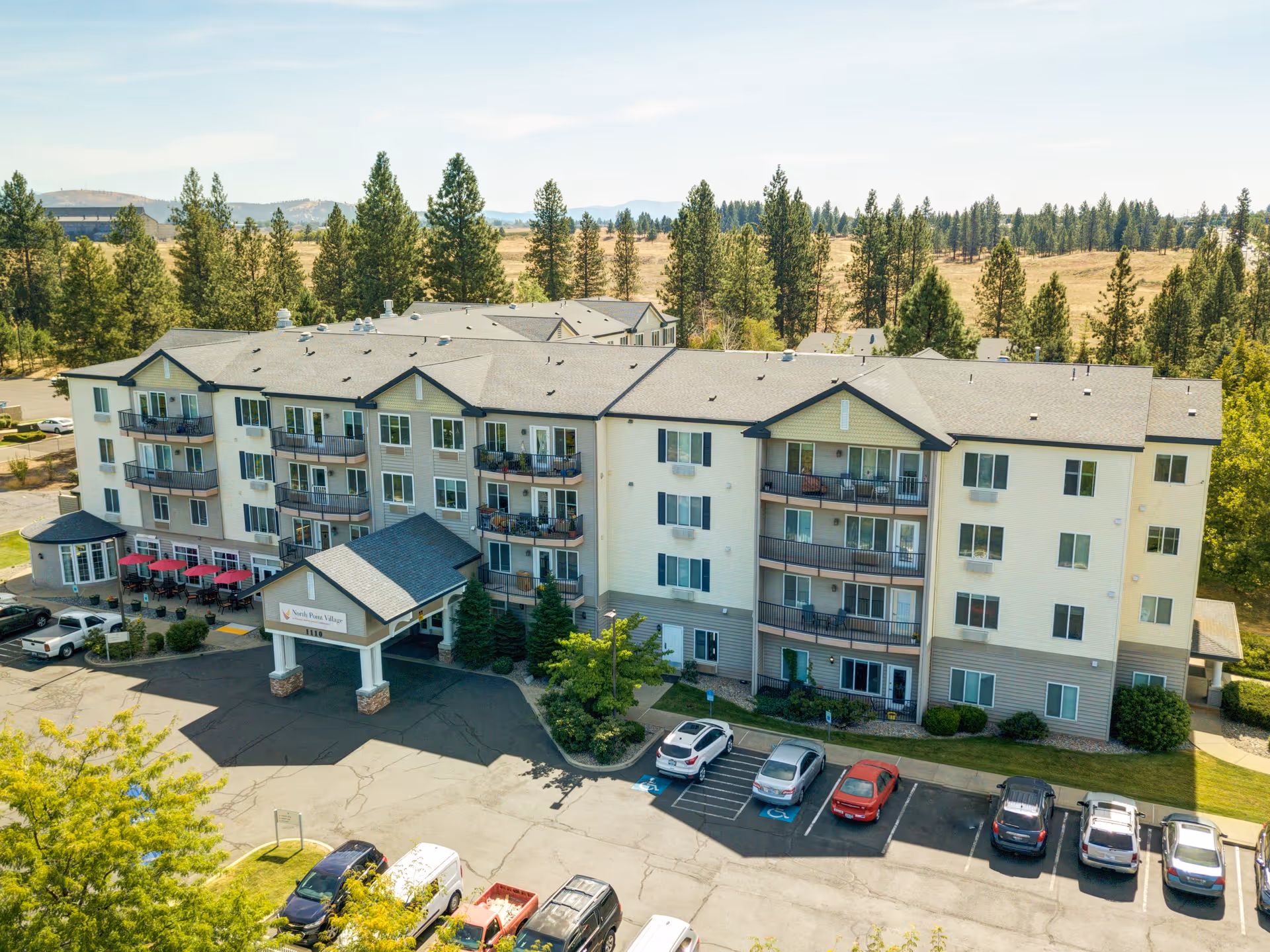 Aerial view of a multi-story senior living facility named North Point Village surrounded by trees and open land, with a parking lot in front containing several cars.