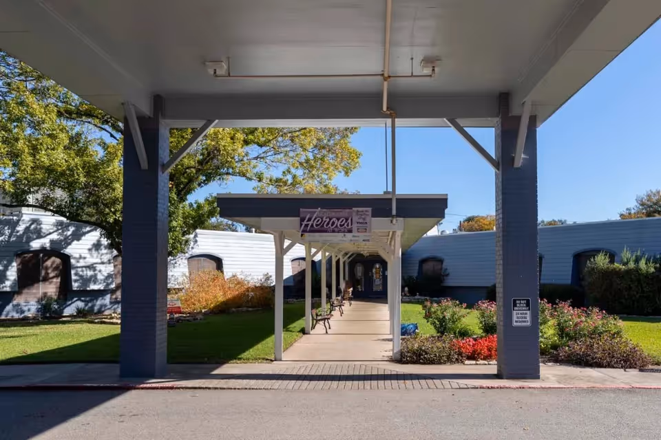 Covered walkway entrance to a building with benches along the path, surrounded by green grass, bushes, and trees under a clear blue sky.