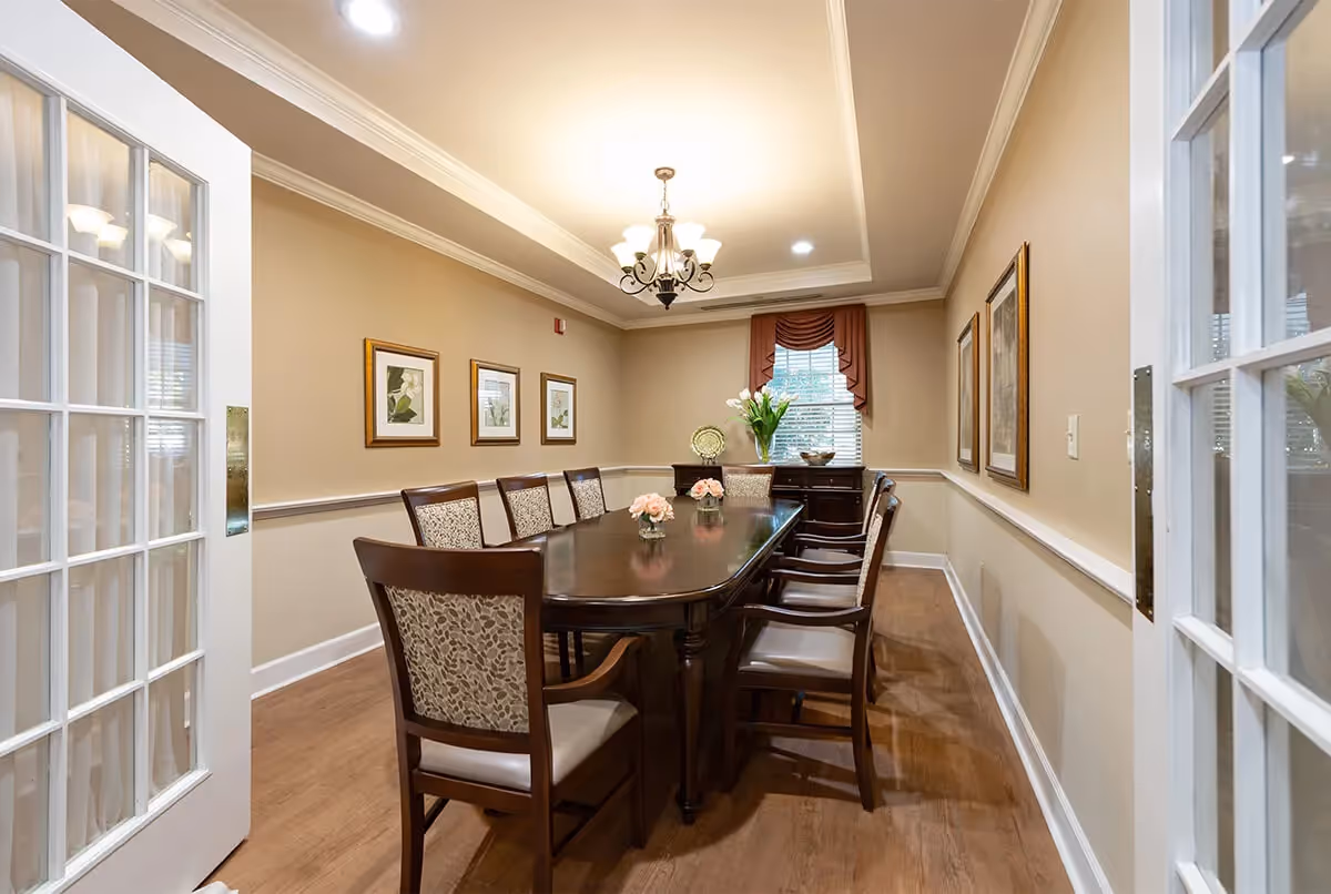 A well-lit dining room with a long wooden table surrounded by eight upholstered chairs. The room features beige walls with white trim, three framed botanical prints on one wall, a window with a red valance, and a chandelier hanging from the ceiling. There is a sideboard with a vase of flowers and decorative items beneath the window. French doors with glass panes are partially open, leading into the room.