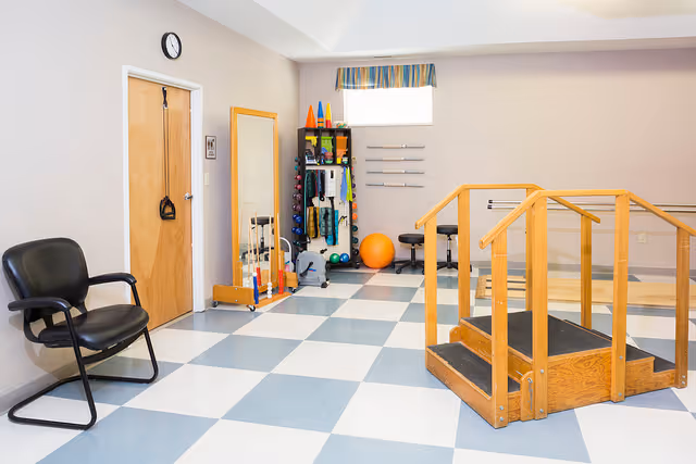 A therapy or rehabilitation room with a black chair, a wooden door with exercise bands hanging on it, a full-length mirror, various exercise equipment including weights, cones, and an exercise ball, and a wooden ramp with handrails. The floor has a blue and white checkered pattern and there is a small window with a striped valance.
