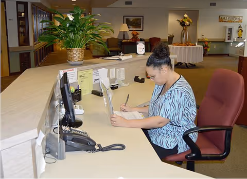 A woman seated at a reception desk in an assisted living center, writing on a clipboard. The desk has a computer monitor, telephone, and a large potted plant. In the background, there are chairs, a table with a flower arrangement, and a sign that reads 'Fall Festival'.