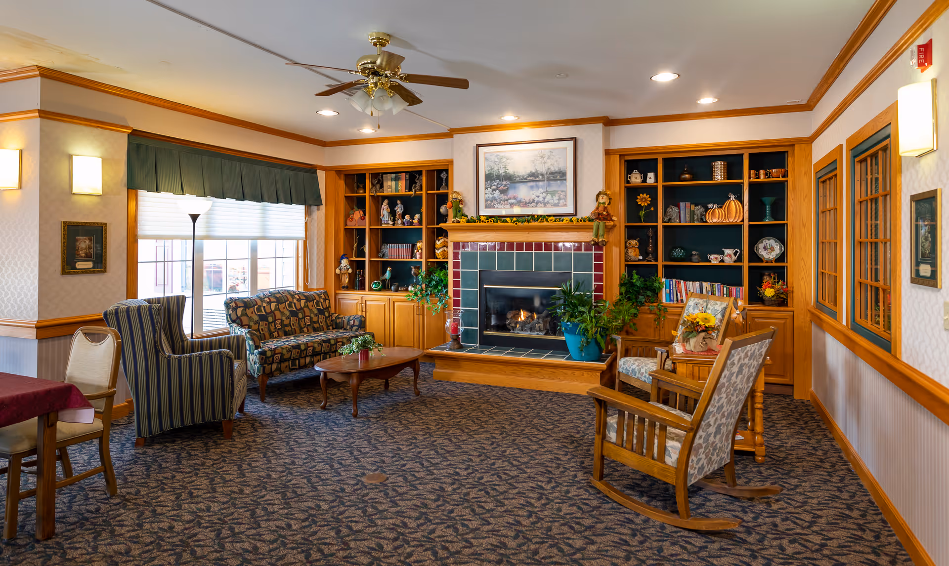 A cozy living room area in an assisted living facility featuring a fireplace with a tiled surround and wooden mantel decorated with plants and figurines. The room has built-in wooden shelves filled with books and decorative items on either side of the fireplace. There are several upholstered chairs and a small sofa arranged around a wooden coffee table. The room is well-lit with ceiling lights, wall sconces, and natural light from a large window with a green valance. The carpet has a patterned design, and the walls are adorned with framed artwork.