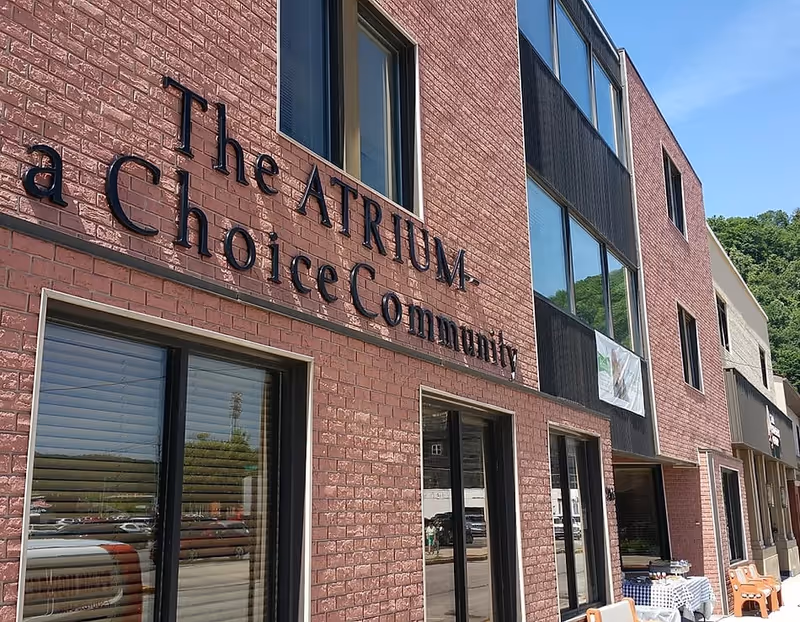 Exterior view of a brick building with large windows and a sign that reads 'The ATRIUM - a Choice Community' on the wall. There are some tables and chairs outside under a clear blue sky.