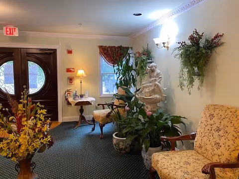 A cozy interior hallway or waiting area with floral-patterned armchairs, several potted plants, a small side table with a lamp, a statue, and a window with a curtain. The area is carpeted and softly lit by wall sconces and natural light from the window. Double wooden doors with circular glass panels are visible at the end of the hallway.