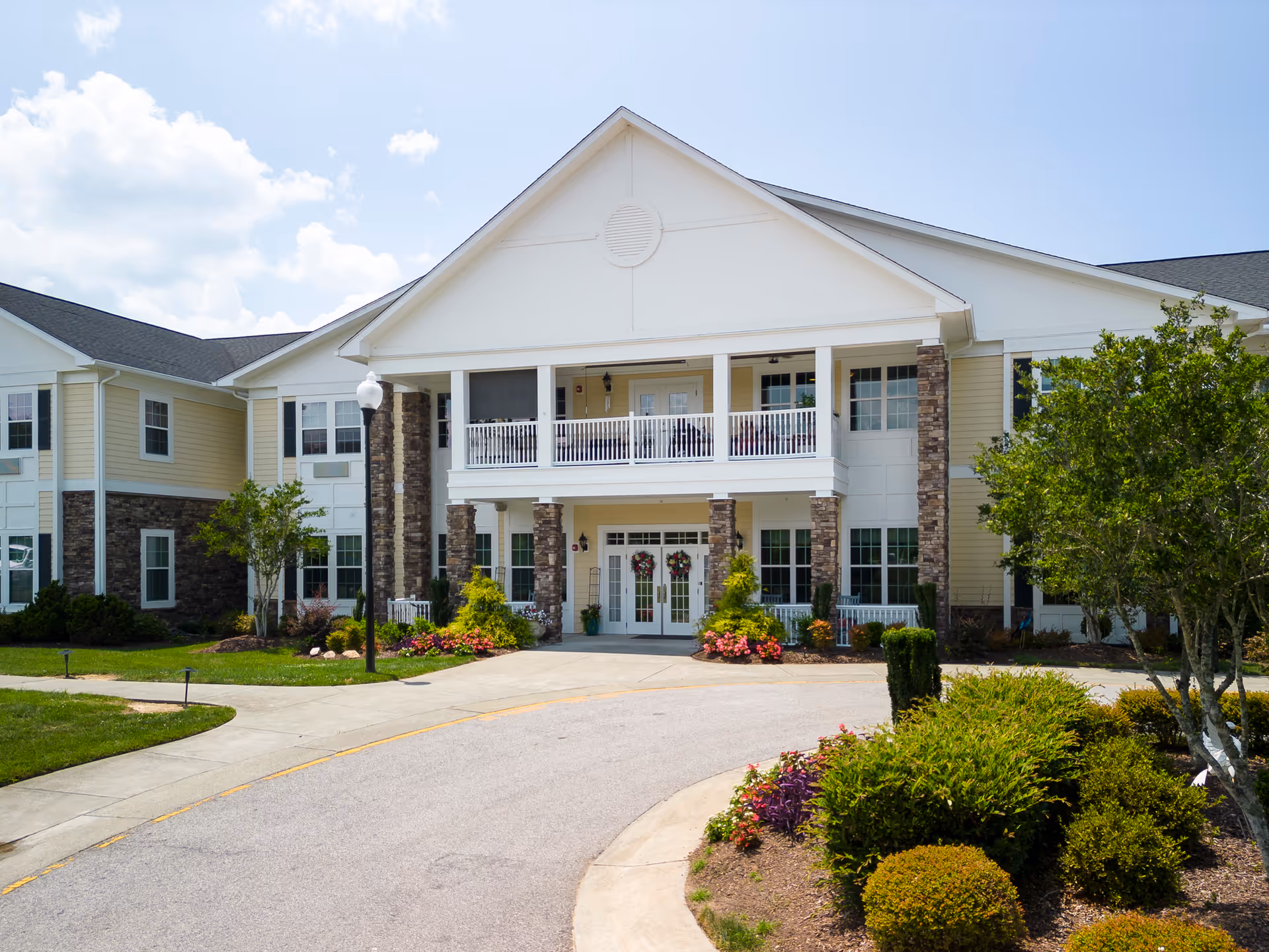 Front exterior view of a two-story senior living facility building with a covered entrance supported by stone pillars, a balcony above the entrance, landscaped bushes and flowers, and a curved driveway leading to the entrance under a partly cloudy sky.