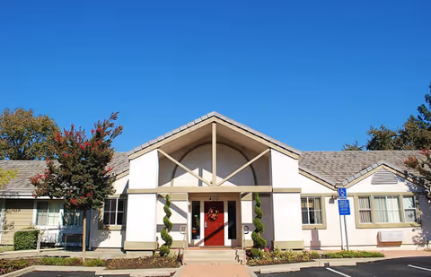 Front exterior of a single-story senior living building with a central gabled entrance, red door, spiral topiary shrubs, and a clear blue sky.