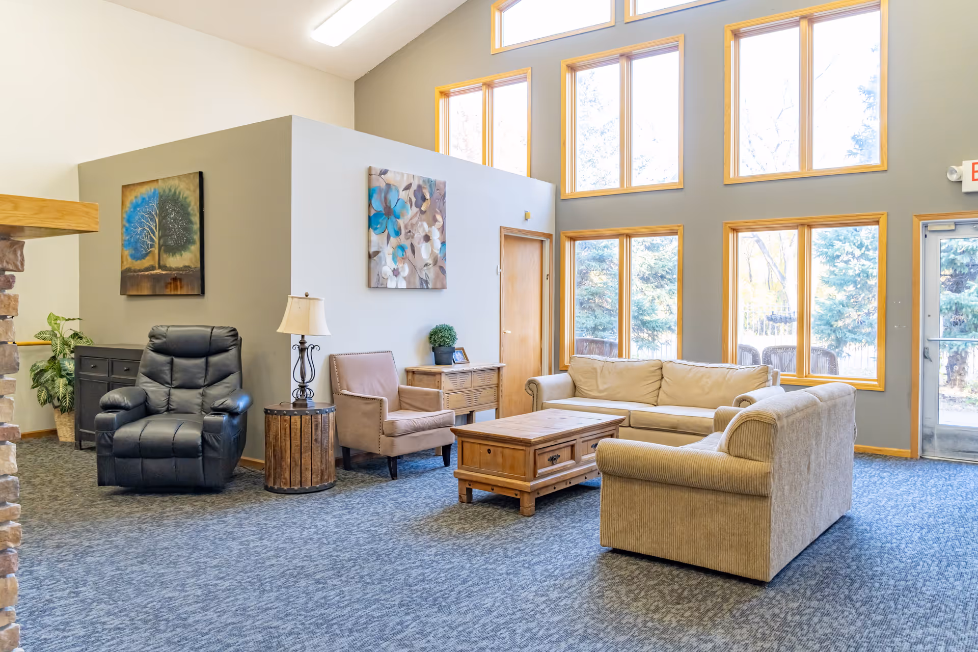 A bright and spacious living room area with large windows letting in natural light. The room features a beige sofa, a beige armchair, a black recliner, a wooden coffee table, a side table with a lamp, and two paintings on the walls. There is a door and a glass exit door visible, with trees outside.