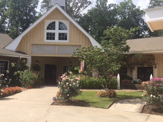 Front exterior view of a building with a peaked roof and large arched window above the entrance. The entrance is surrounded by well-maintained flower beds with blooming roses and other plants. There is a concrete driveway and walkway leading to the entrance, with trees and greenery around the building.