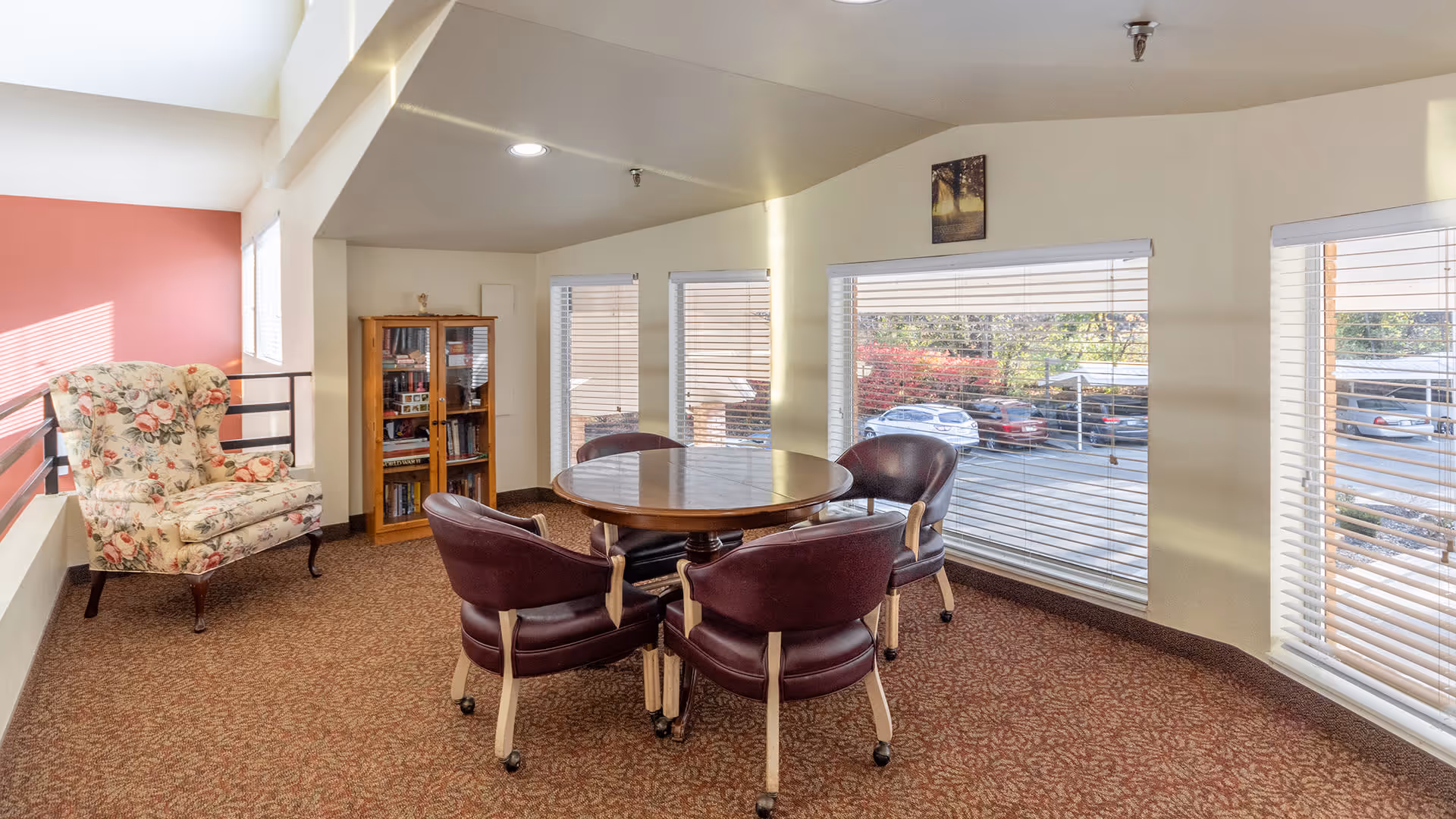 A cozy sitting area with a round wooden table surrounded by four maroon cushioned chairs on wheels. To the left, there is a floral upholstered armchair next to a wooden bookshelf filled with books. Large windows with white blinds let in natural light and offer a view of parked cars outside. The walls are painted light yellow with one accent wall in a muted red tone.