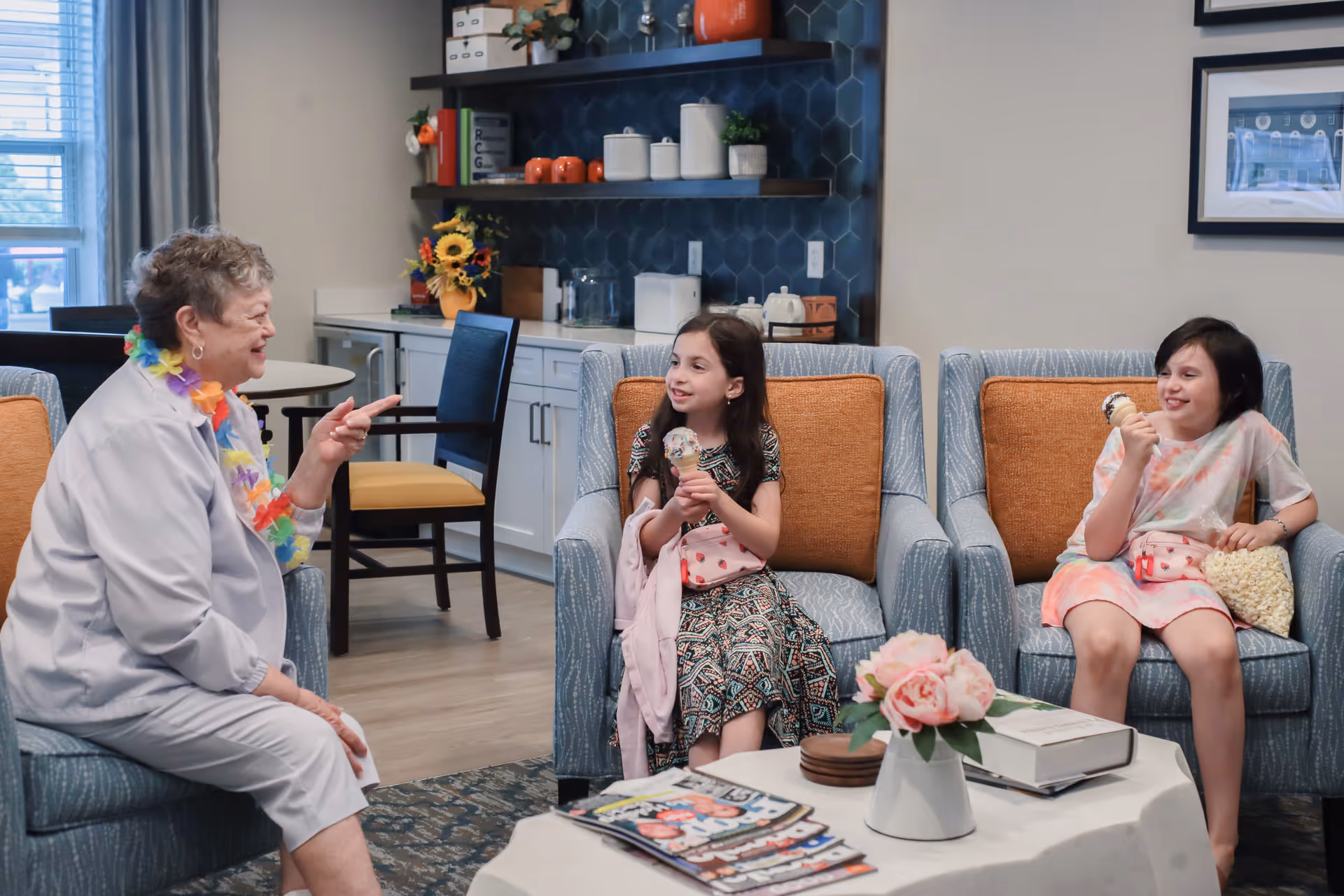 An elderly woman chats with two young girls eating ice cream while seated in a comfortable common living room area.