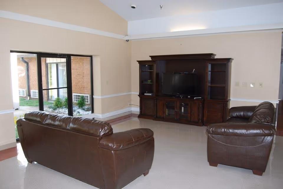 Communal living room with two brown leather sofas facing a wooden entertainment center and television, with large windows looking out to a courtyard.