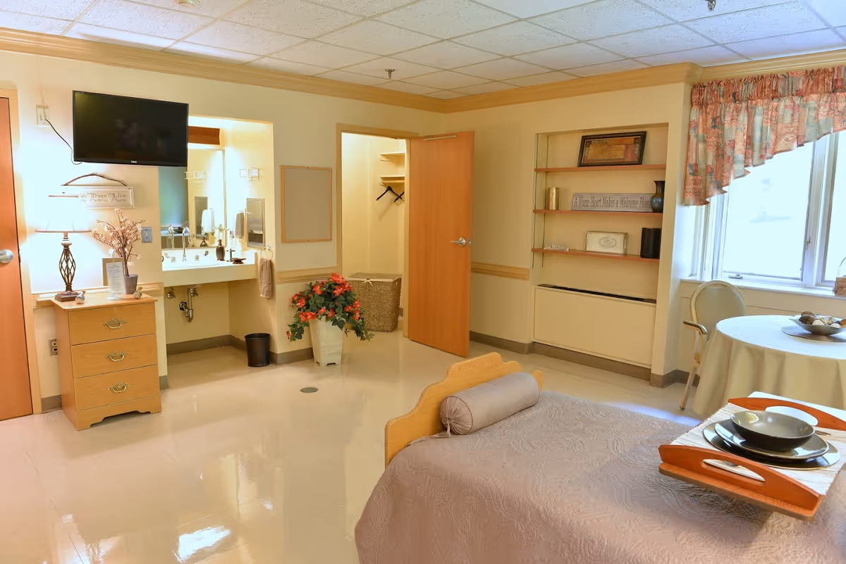 A well-lit bedroom in a senior living facility featuring a single bed with a bolster pillow and a tray with dishes on it. There is a small wooden dresser with a lamp and decorative items, a wall-mounted TV, an open closet with hangers, a sink area with a mirror, a potted plant, built-in shelves with framed pictures and decor, and a round table with chairs near a window with floral curtains.