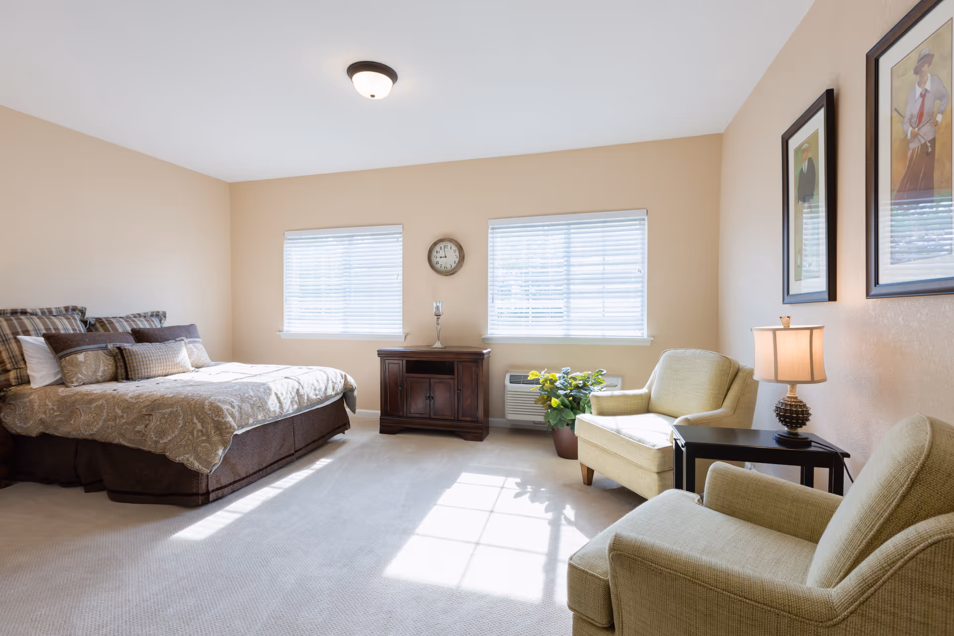 A bright and spacious bedroom with a large bed on the left side, two windows with blinds letting in natural light, a wooden cabinet with a clock and candle holder between the windows, and two beige armchairs with a small black side table and lamp on the right side. Two framed pictures hang on the wall above the armchairs.