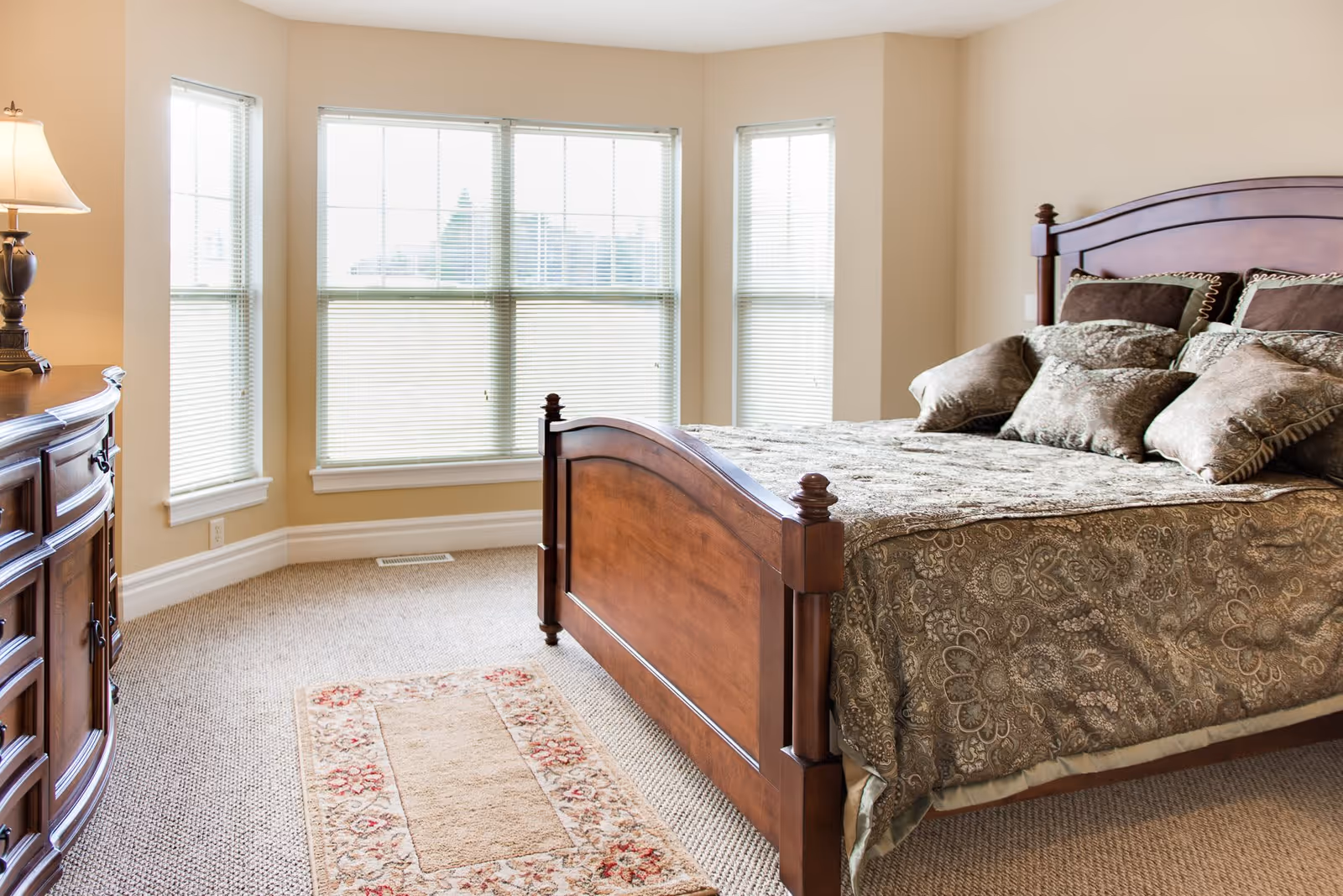 A bright bedroom with a large wooden bed featuring a patterned bedspread and multiple pillows. The room has beige walls, a carpeted floor, a wooden dresser with a lamp on it, and three large windows with blinds allowing natural light to fill the space.