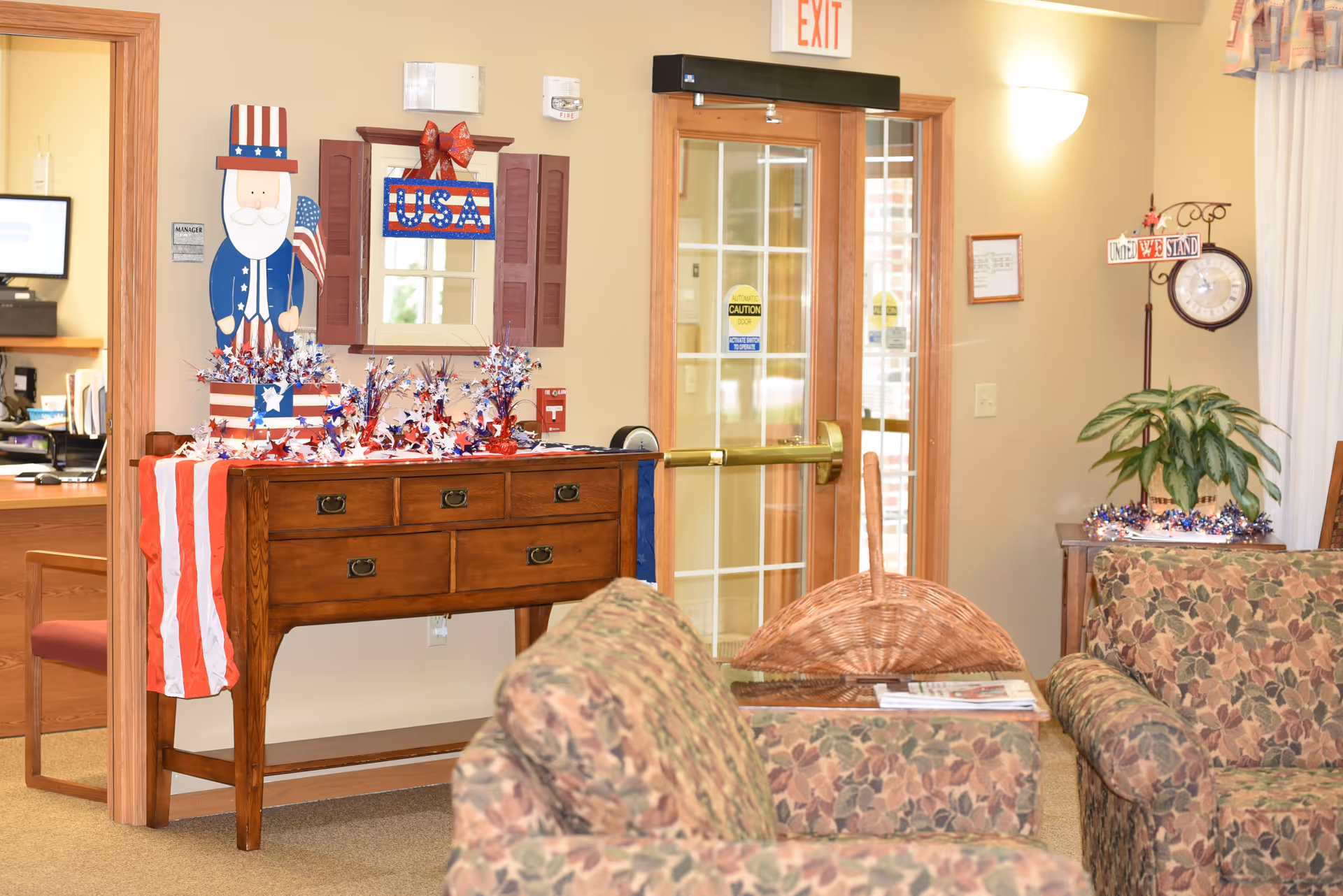 A cozy interior area with floral-patterned armchairs, a wooden console table decorated with patriotic USA-themed decorations including a wooden Uncle Sam figure, red, white, and blue stars, and a USA sign. There is a glass door with an exit sign above it, a clock on a stand with a sign that reads 'United We Stand,' and a potted plant on a small table. The area appears well-lit and welcoming.