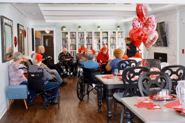 A group of elderly people, some in wheelchairs, gathered in a bright room with bookshelves along the back wall. The room has wooden floors, a fireplace, and tables decorated with red Valentine's Day themed items and balloons. Several people are wearing red hats and festive attire.