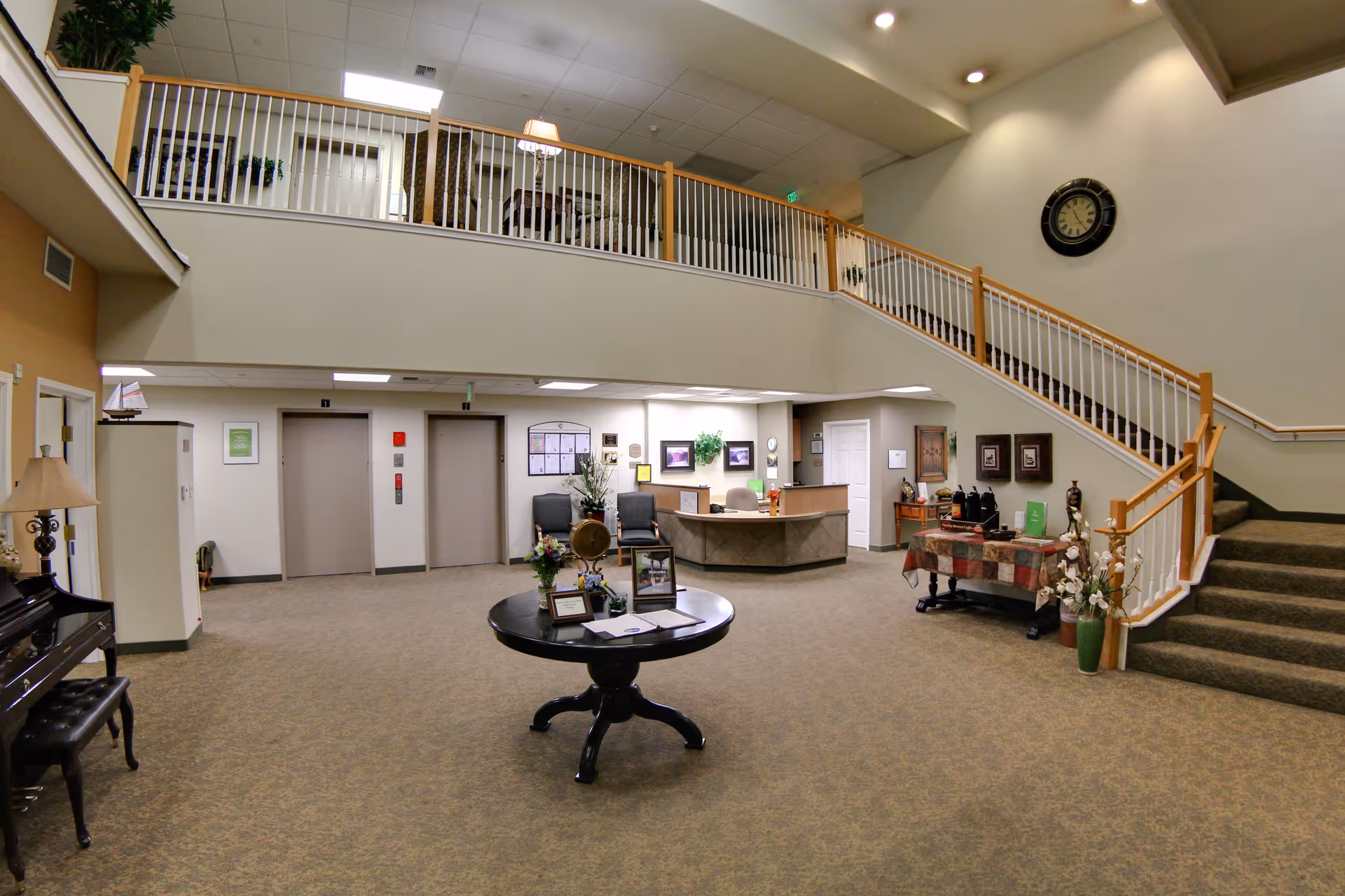 Spacious two-story senior living facility lobby with a reception desk, central round table, seating, and a staircase to the upper level.