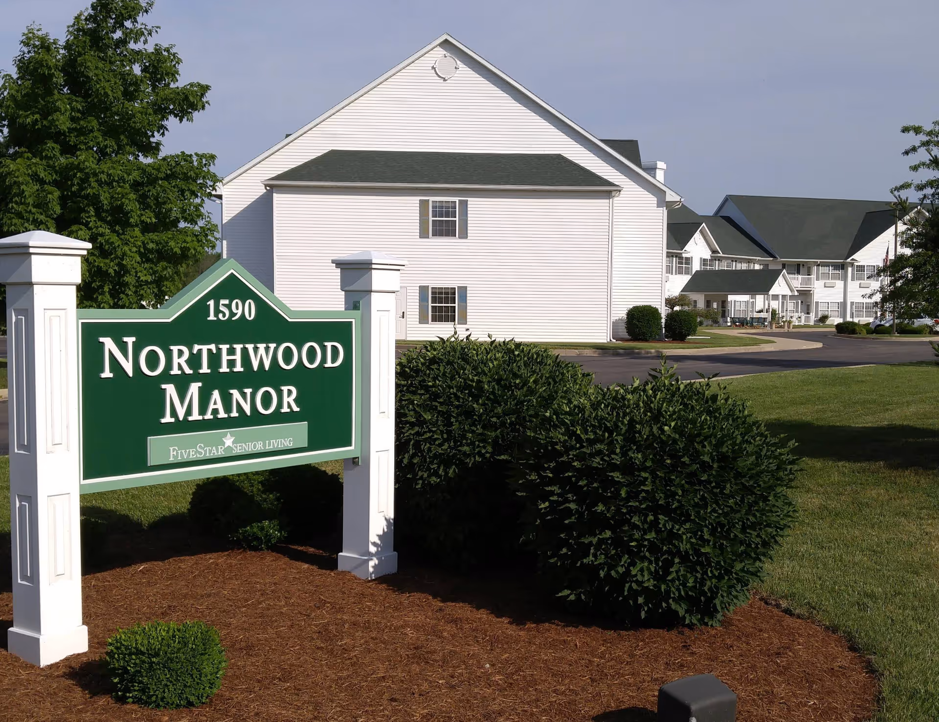 Exterior view of Northwood Manor senior living facility with a green and white sign displaying the name and address 1590. The building is white with green roofs and surrounded by well-maintained bushes and lawn.