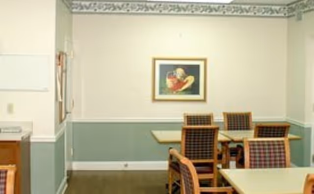 A dining area with several tables and chairs featuring plaid upholstery. The walls are painted in a two-tone style with a decorative border near the ceiling. A framed painting hangs on the wall.