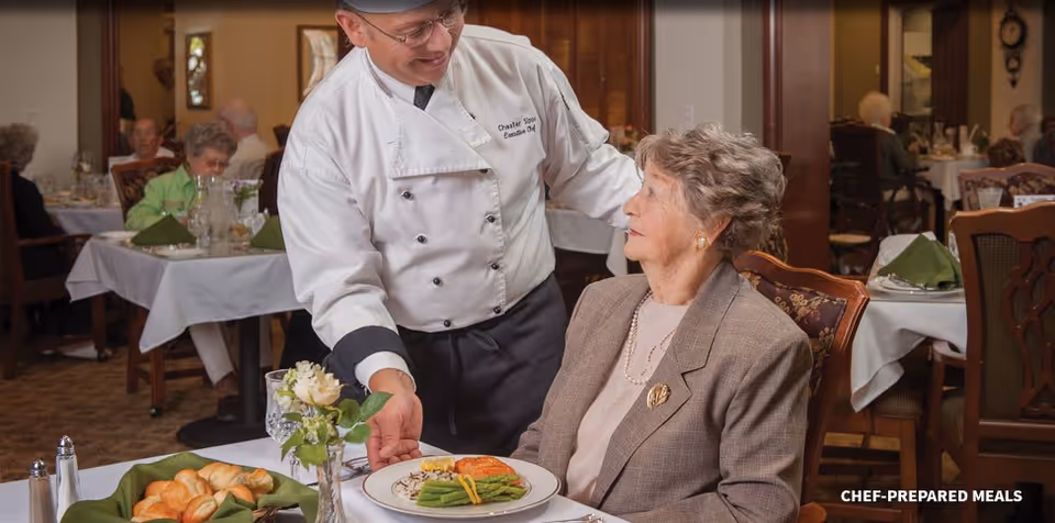 A chef serves a plated meal to an elderly woman seated at a table in a communal dining room.