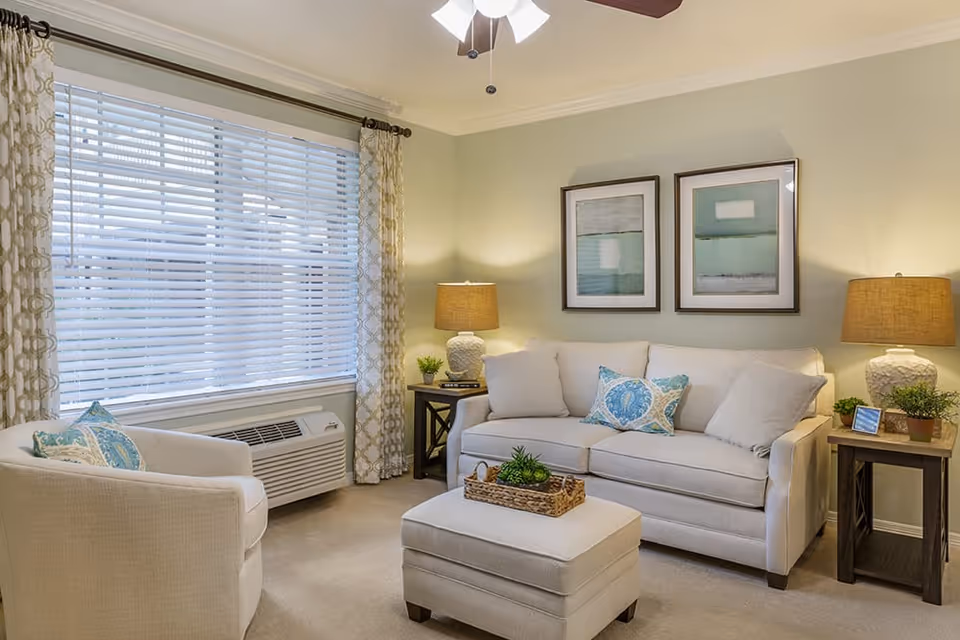 A cozy living room with a beige sofa adorned with decorative pillows, a matching armchair, and an ottoman with a wicker tray holding a small plant. Two framed abstract artworks hang on the light green wall above the sofa. There are two side tables with table lamps and small plants, a large window with white blinds and patterned curtains, and a ceiling fan with lights.