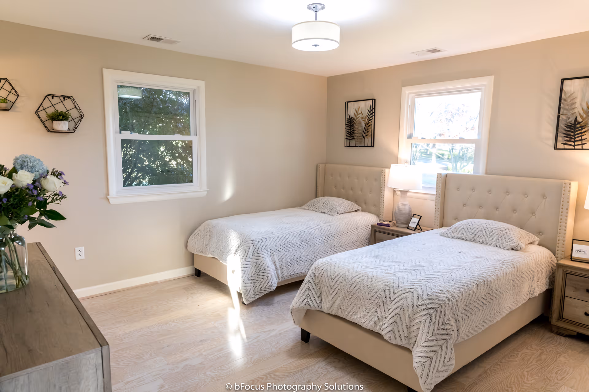 A bright bedroom with two twin beds featuring beige tufted headboards and white patterned bedspreads. There are two windows letting in natural light, two bedside tables with lamps, framed botanical artwork on the walls, a wooden dresser with a vase of flowers, and light wood flooring.
