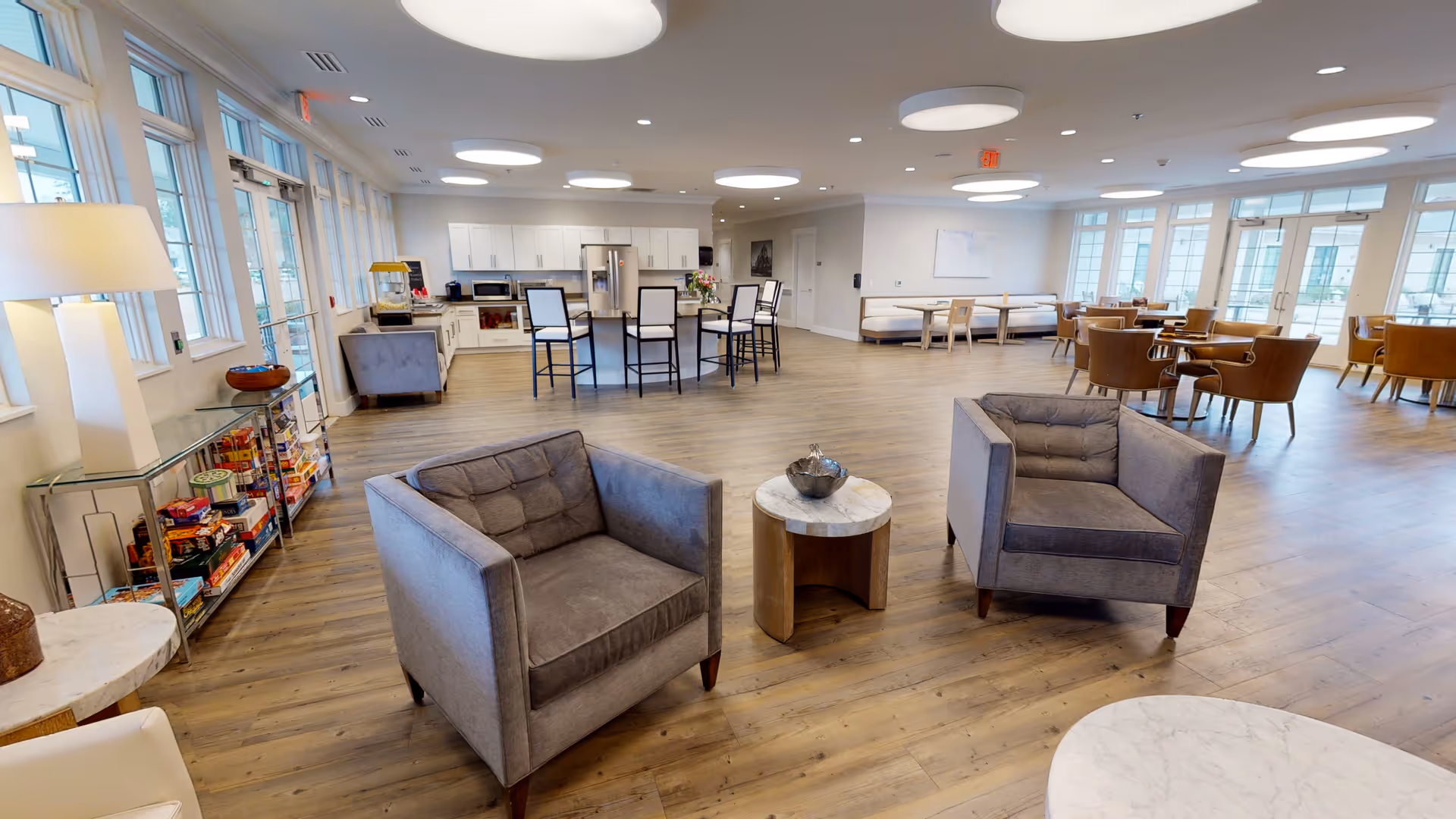 A spacious, well-lit common area in a senior living facility featuring two gray armchairs around a small round table in the foreground, a kitchen area with white cabinets and a central island with high chairs in the background, and multiple seating areas with tables and chairs near large windows letting in natural light.