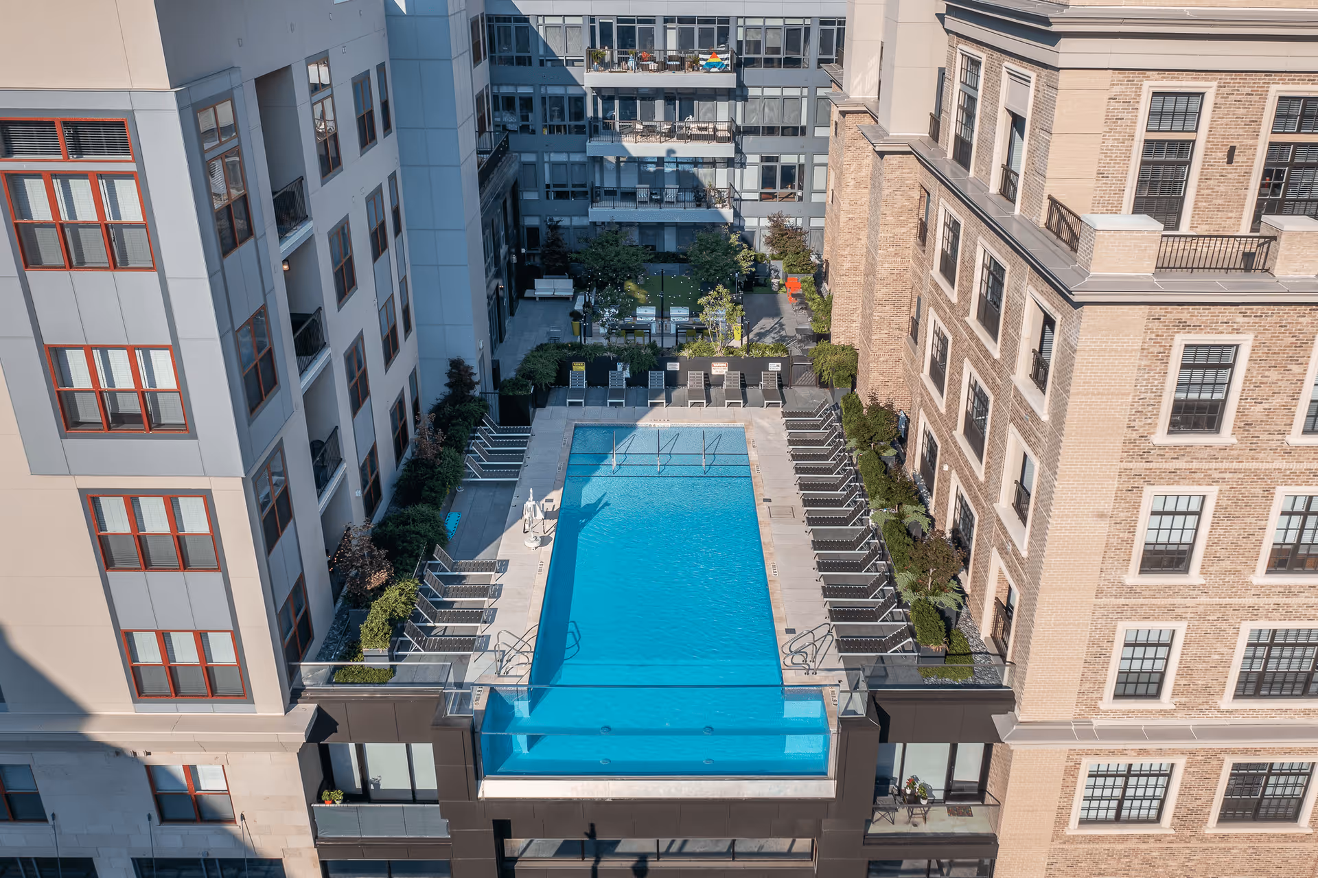 A rooftop swimming pool with clear blue water surrounded by numerous lounge chairs. The pool is situated between two multi-story buildings with windows and balconies. There are plants and greenery around the pool area, and a small garden space with seating and tables is visible beyond the pool.